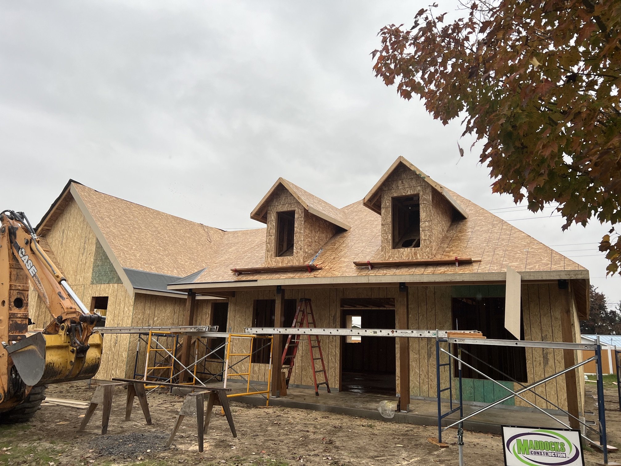 Construction of a house with wooden frame and roof, scaffolding, ladders, and construction equipment present at the site, under a cloudy sky.