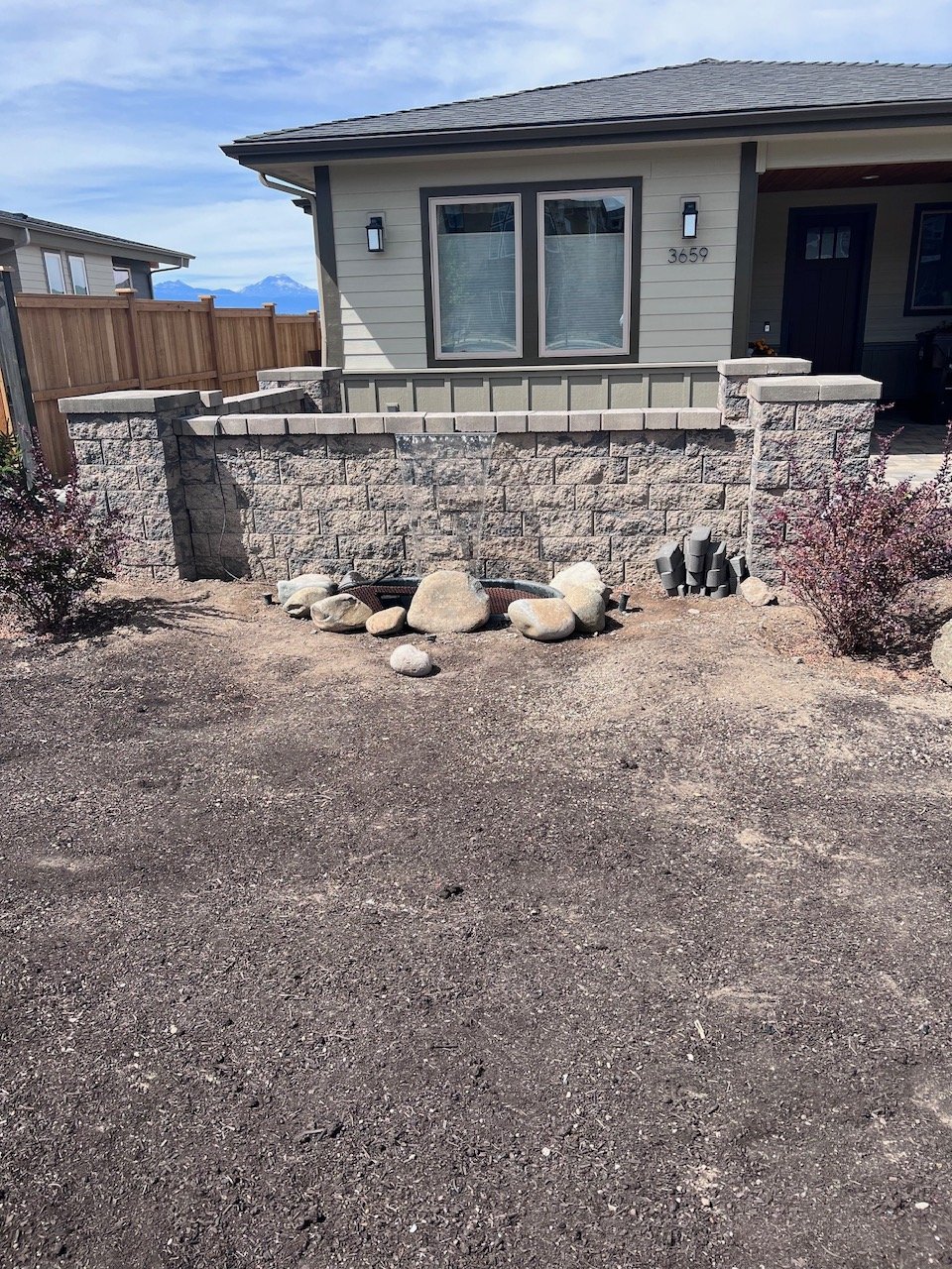 Front yard with a stone retaining wall, rocks, bushes, and the house number 3659 on a light-colored house, with mountains visible in the background.