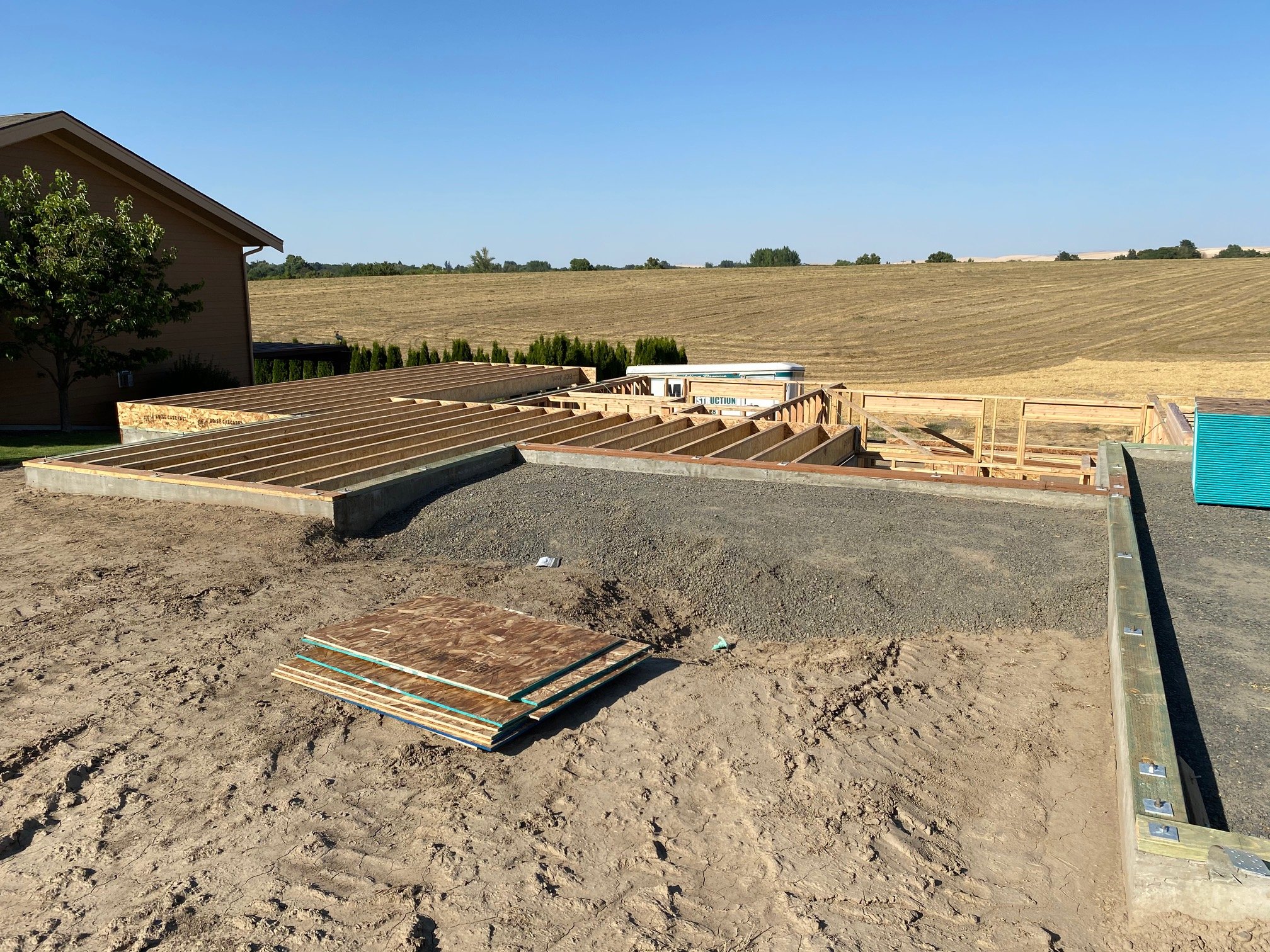 Construction site with a wooden foundation frame for a building, surrounded by dirt and a backyard with a house and a tree, with open fields and a clear blue sky in the background.