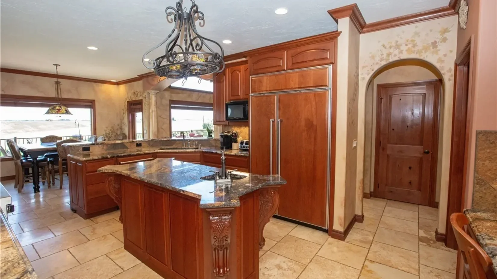Kitchen with wooden cabinets, a granite countertop island, and a dining area with large windows showing an outdoor view.
