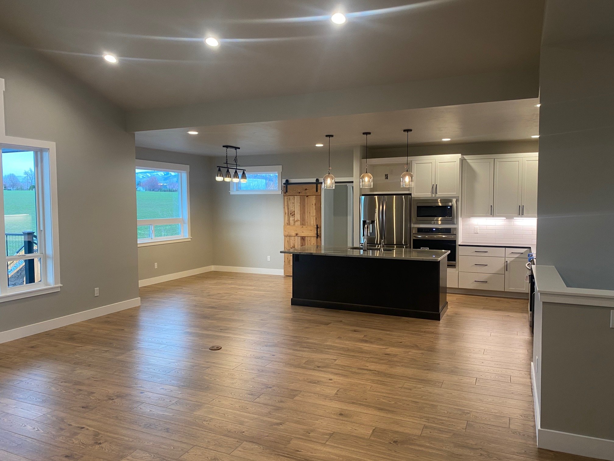 View of a modern, open-concept kitchen with white cabinets, stainless steel appliances, a black kitchen island, pendant lights over the island, and large windows showing an outdoor landscape with grass and trees.