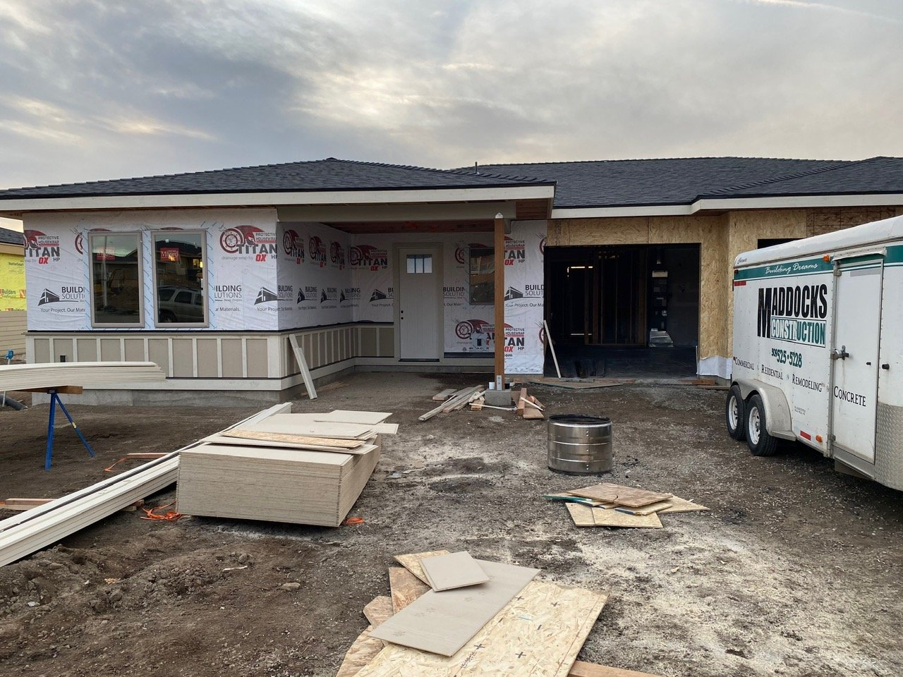 Under construction house with partially completed walls, construction materials, and a trailer on a dirt lot.