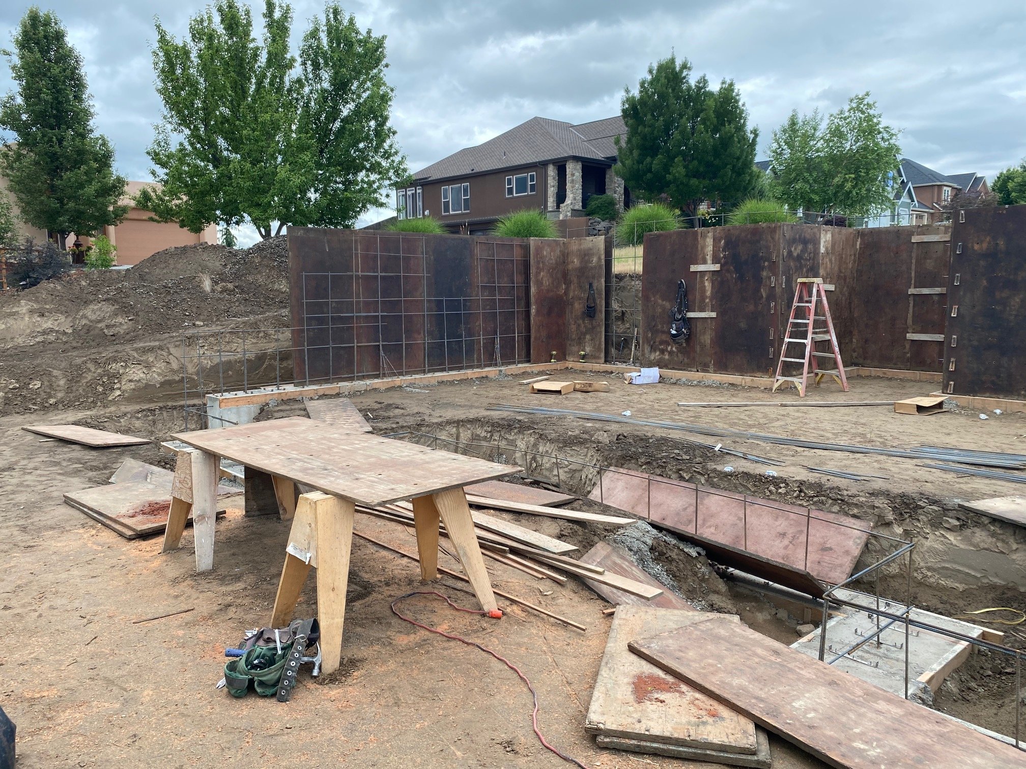 Construction site with excavation area and wooden formwork for a building foundation, construction tools, and a ladder, with houses and trees in the background under cloudy sky.