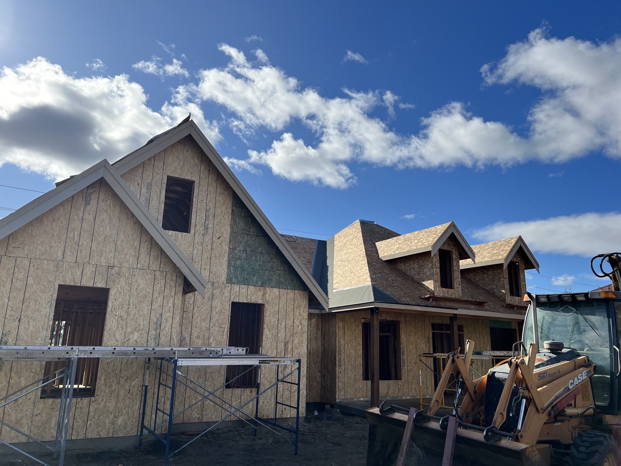 Construction site with unfinished houses covered in plywood, scaffolding, construction equipment, and a bright blue sky with scattered clouds.