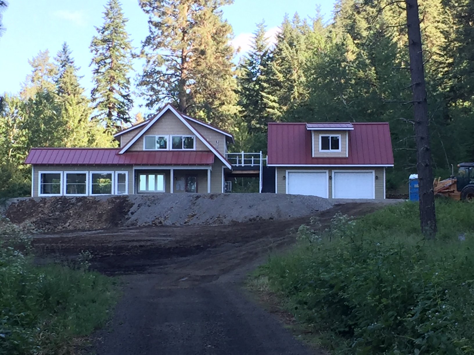 A house under construction in a wooded area, with a dirt road leading up to it. The house has a red metal roof, large windows, and two garage doors.