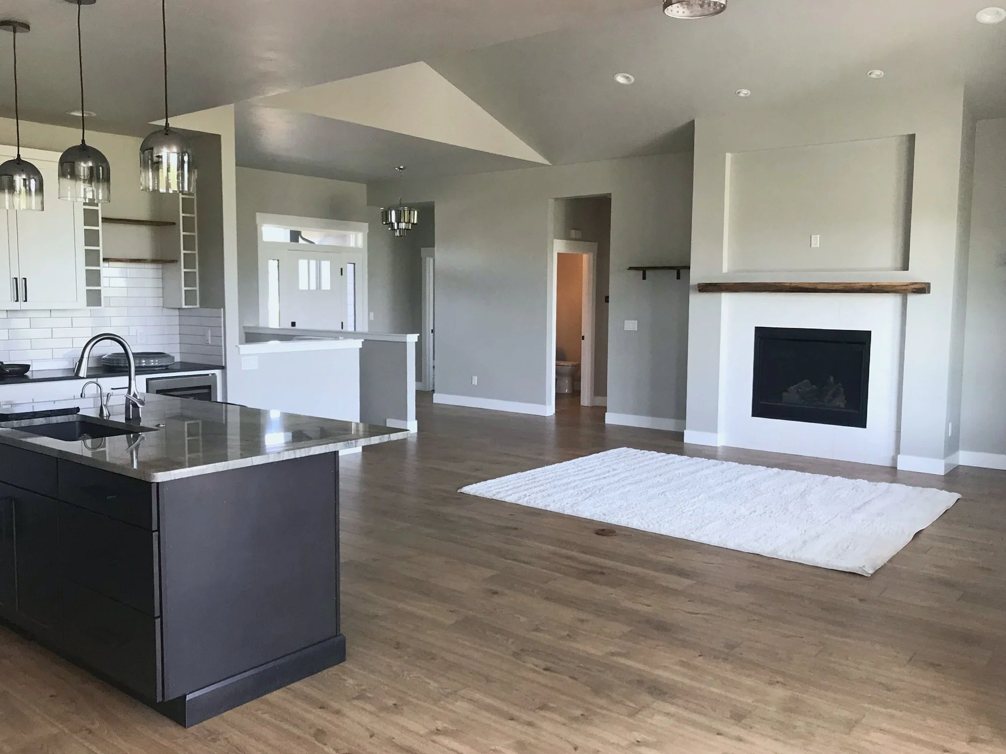 Open-concept modern kitchen with an island, black cabinets, pendant lighting, and a view into a living area with a fireplace and white rug.