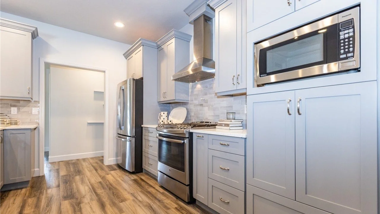 Modern kitchen with light gray cabinets, stainless steel refrigerator, oven, microwave, and hood, wood flooring, and a stack of books on the counter.