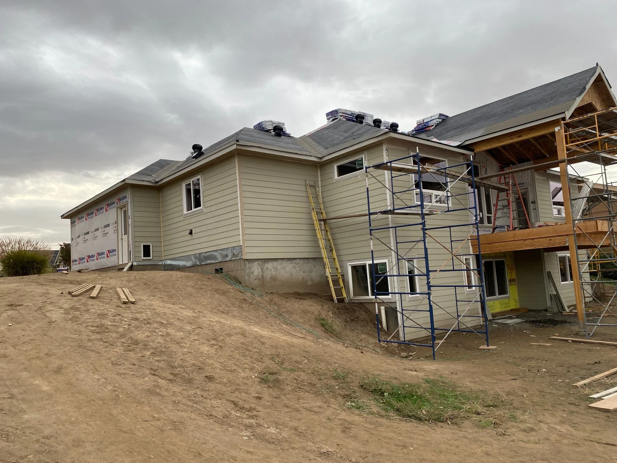 A two-story house under construction with siding being installed. The roof is unfinished with visible shingles on one side. There is scaffolding and ladders around the house, and construction materials are stacked on the roof. The sky is overcast.