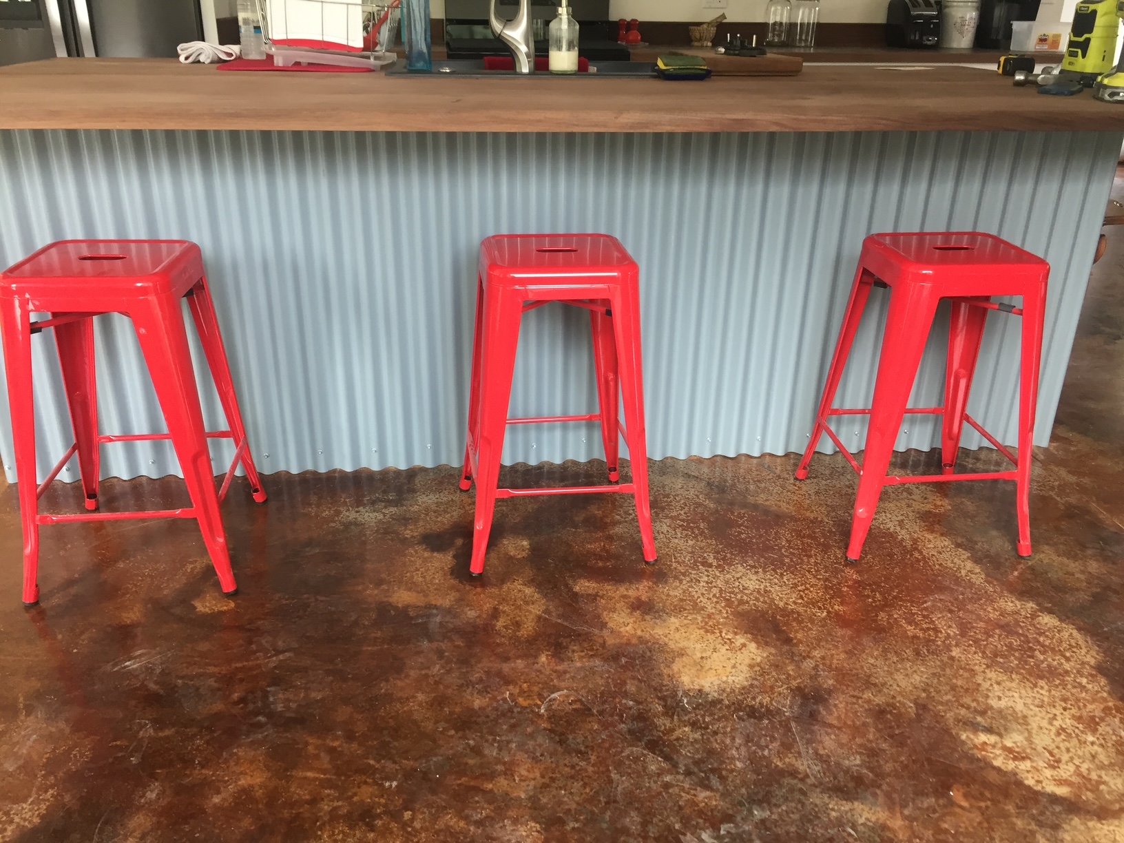 Three red metal barstools positioned in front of a kitchen counter with a wood tabletop and a metal panel front. The counter has various kitchen items and appliances on it.