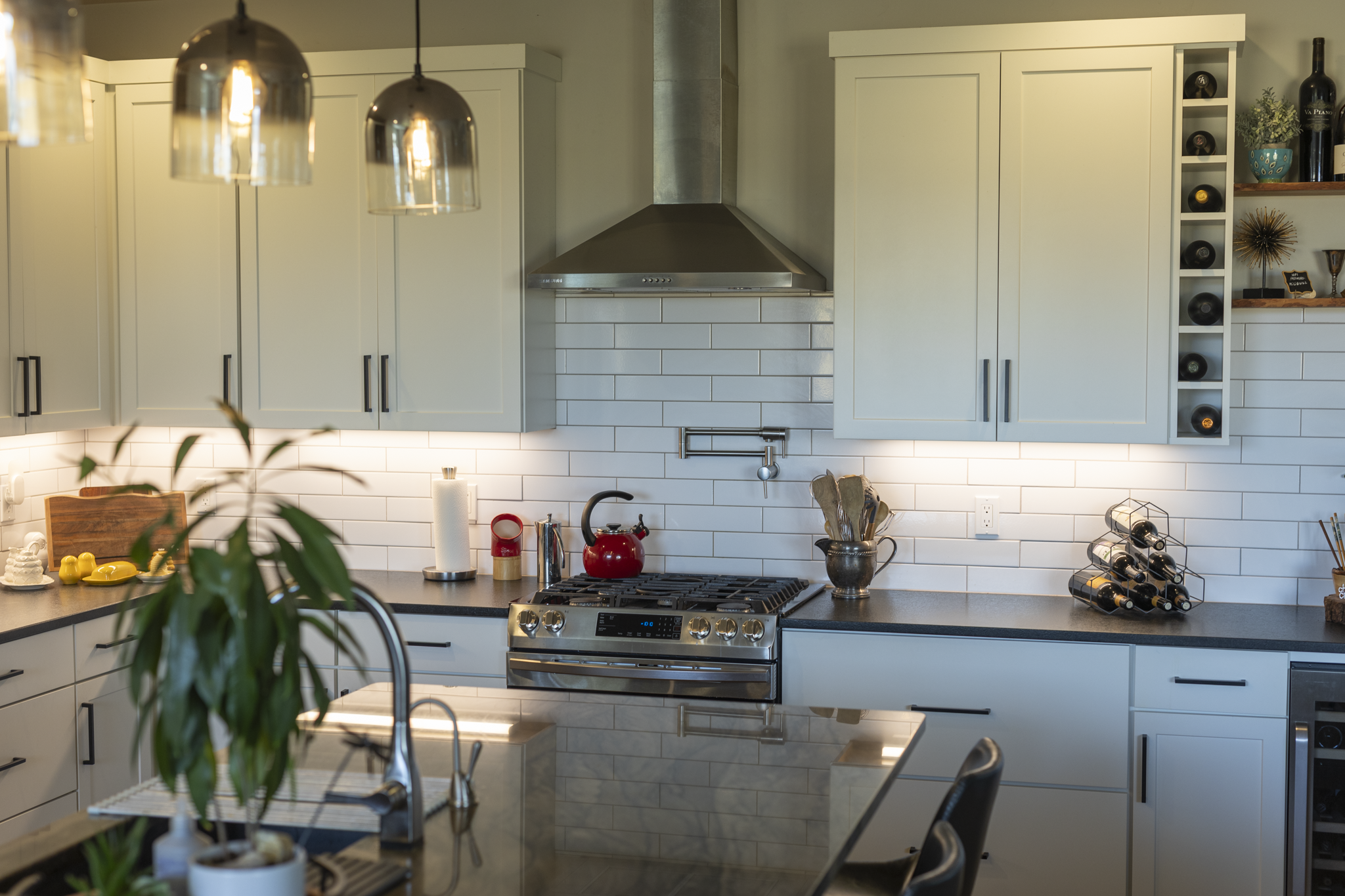 A modern kitchen with white cabinetry, a stainless steel range hood, a black countertop, and decorative items including wine bottles, a plant, and kitchen utensils.