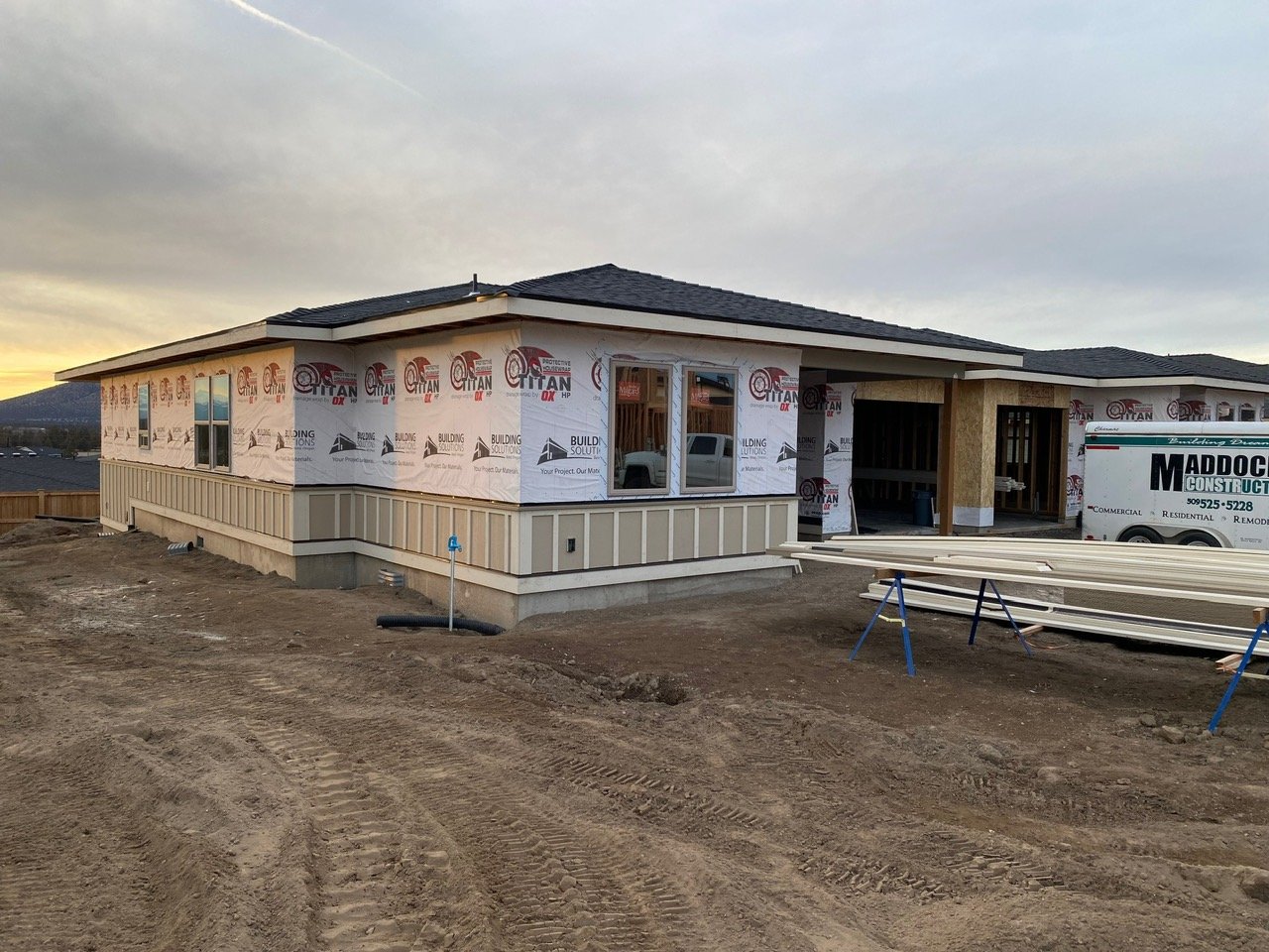 Construction site of a house with new siding and windows, unfinished structures, and construction materials on the ground at sunset.