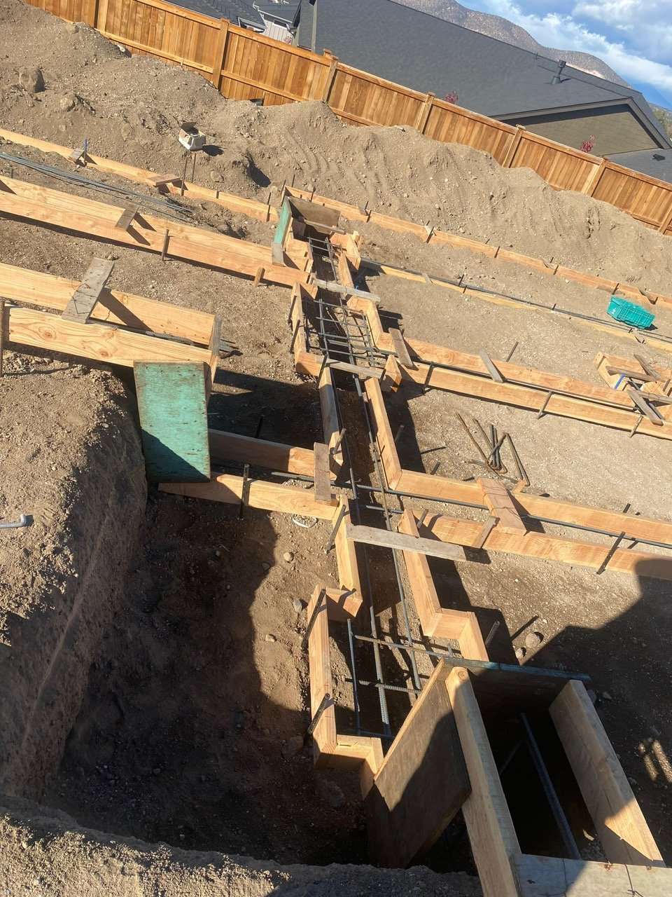 Construction site with wooden formwork and rebar for building the foundation, surrounded by dirt and partially excavated hole.