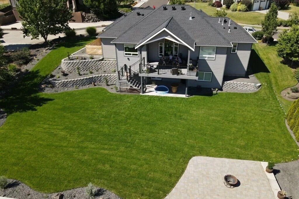 A two-story gray house with a backyard featuring a well-maintained green lawn, a patio with outdoor furniture, and landscaped flower beds on a sunny day.