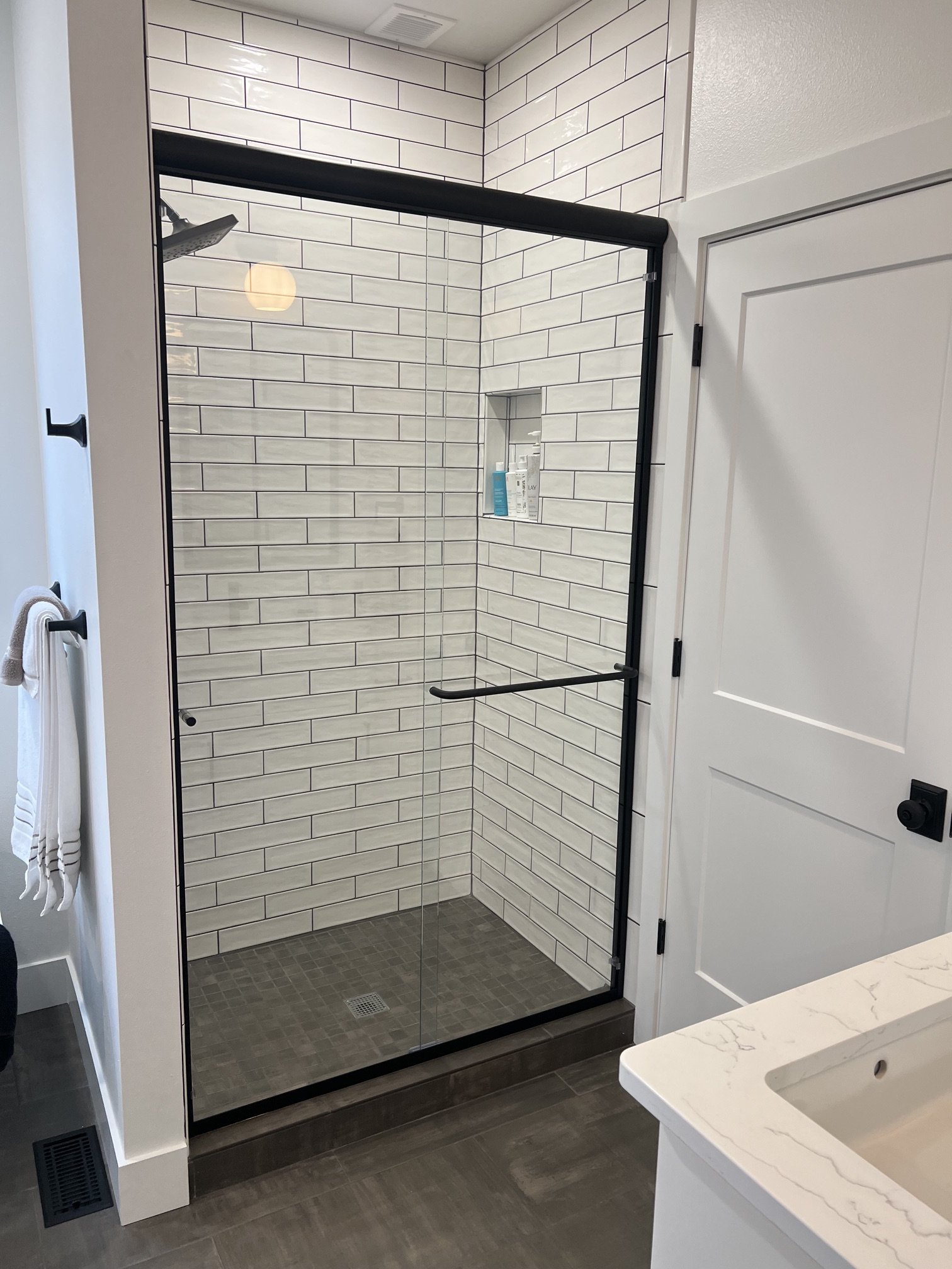 A bathroom with a walk-in shower enclosed by glass with black frame, white subway tile walls, and small dark floor tiles. Part of a white vanity with marble countertop is visible in the foreground, with wall towels on hooks on the left side of the im
