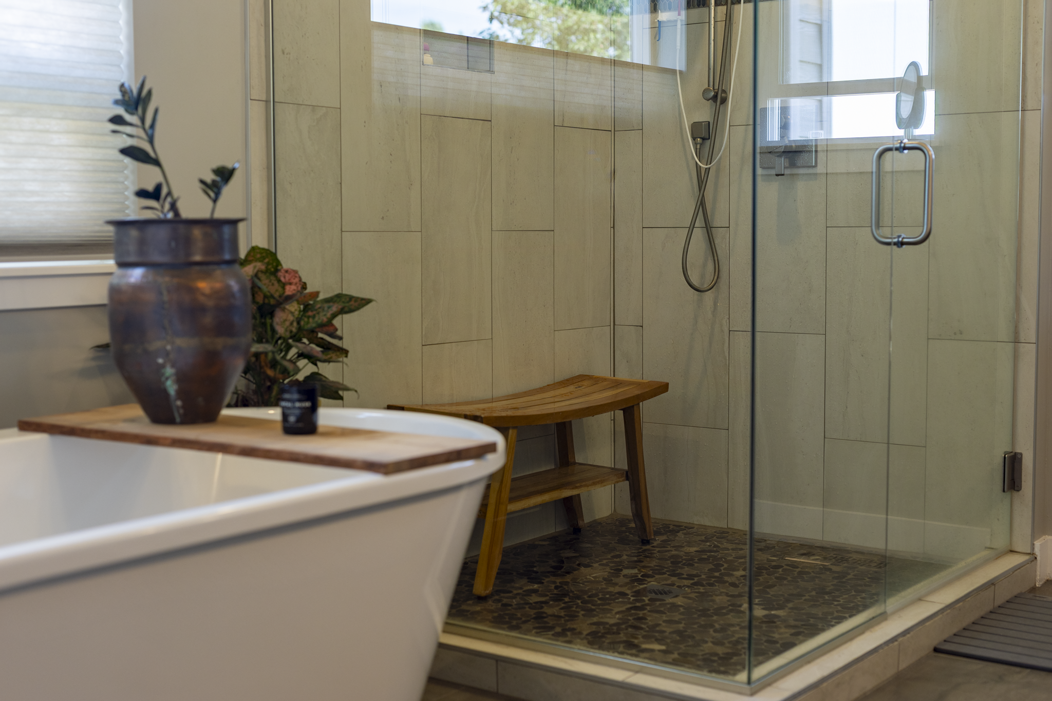 A modern bathroom with a glass-enclosed shower, beige tiled walls, a wooden bench, and a white bathtub with a wooden tray holding a potted plant and a black container. Natural light streams in through a window with blinds.