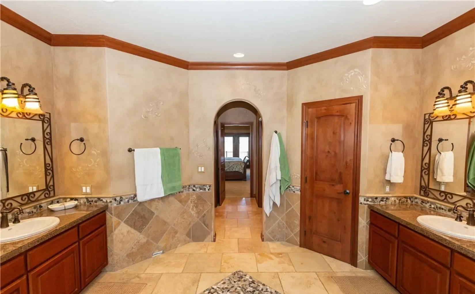 A spacious bathroom with two sinks and large mirrors, beige tiled walls with decorative border, wooden cabinets, and towels hanging on black metal towel racks. An archway leads to a bedroom with a bed and window.