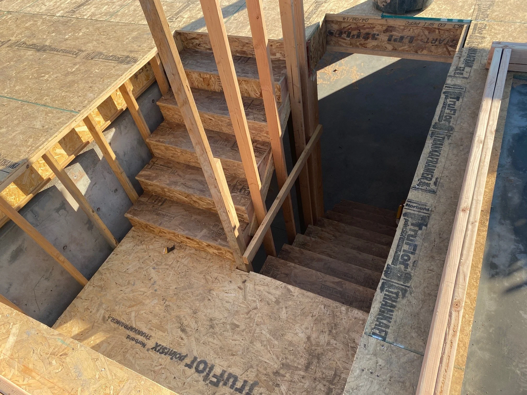 Construction site featuring a partially built staircase with wooden framing and plywood sheathing, surrounded by construction materials and unfinished flooring.