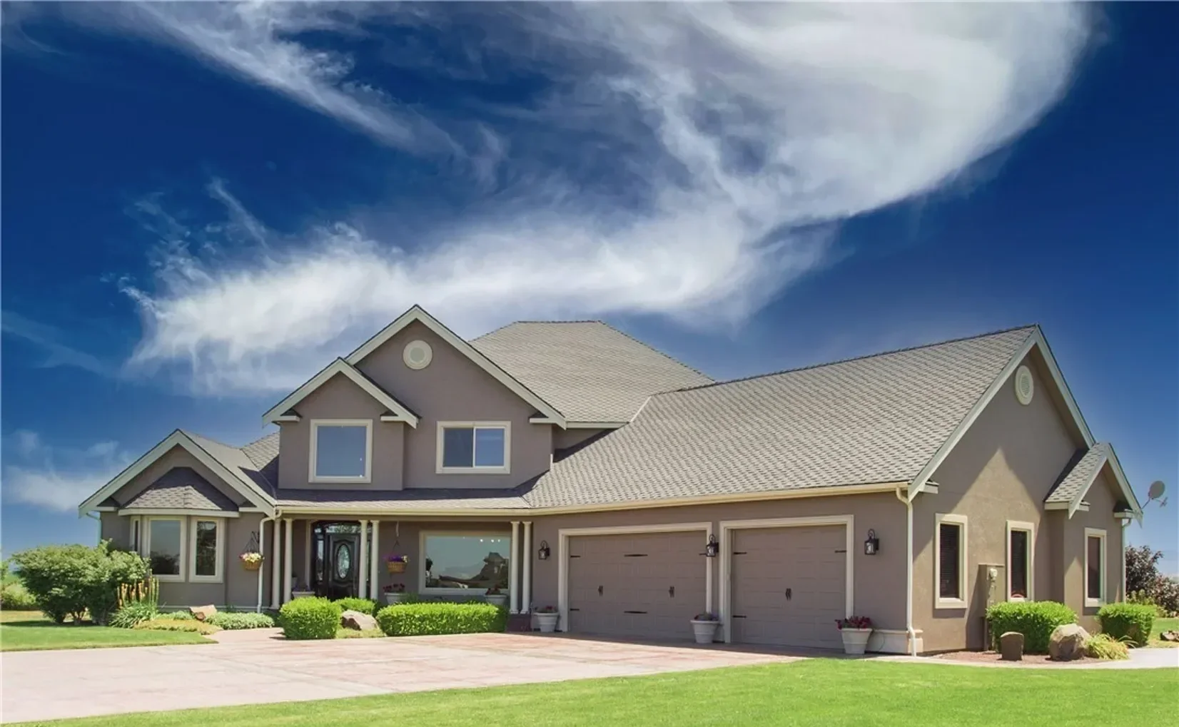 A modern two-story house with beige siding, gray roof, front lawn, and driveway under a partly cloudy blue sky.