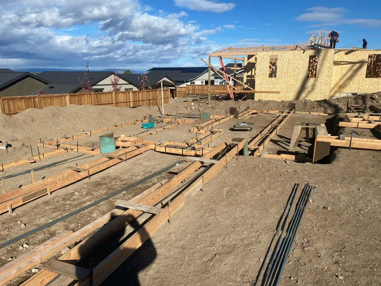 Construction site with wooden framing for a house in progress, with workers on top and a residential neighborhood in the background under partly cloudy skies.