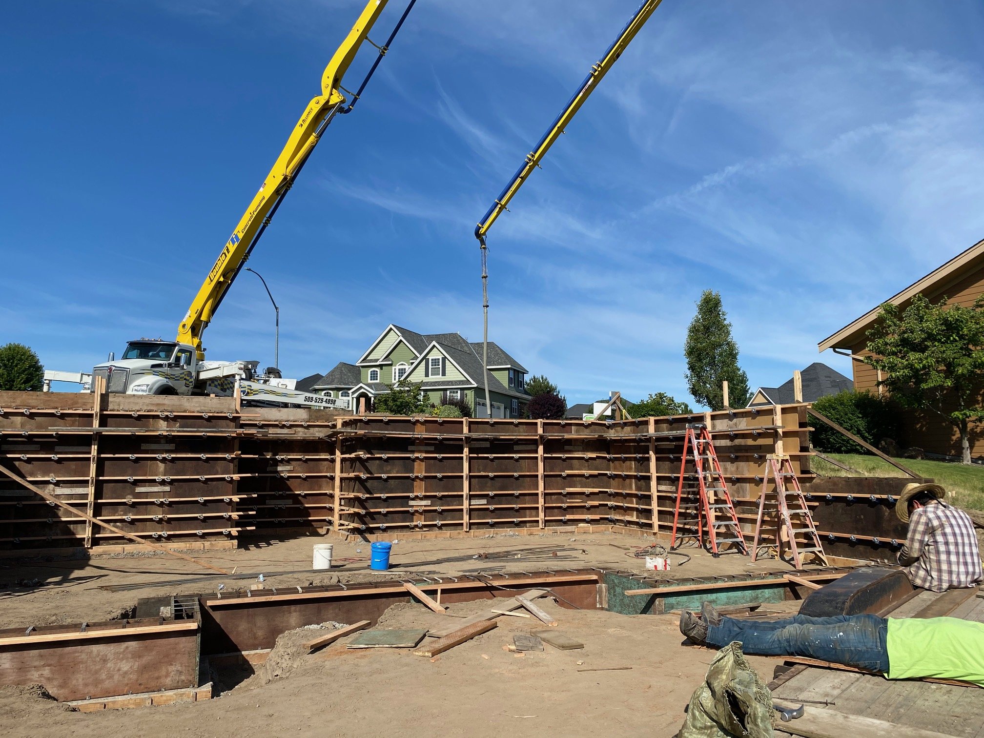 Construction workers building a house foundation with wooden forms and steel rebar, using a crane to set the concrete wall, in a neighborhood with houses and trees under a blue sky.