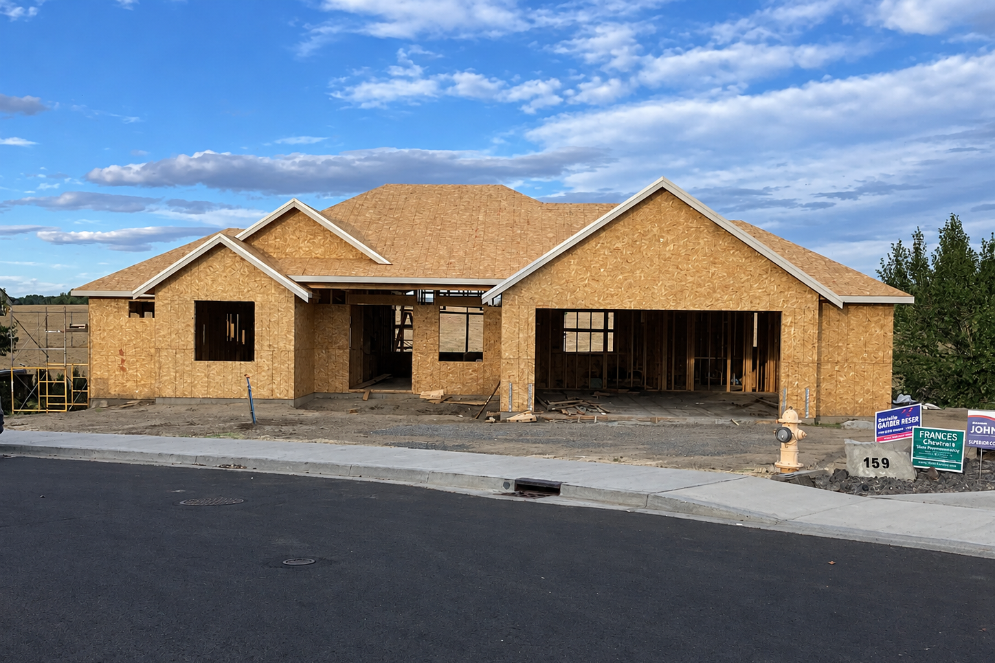A house under construction with unfinished wooden walls and a partially completed roof. The house has no windows or doors installed yet and is situated next to a paved road with an orange fire hydrant nearby.