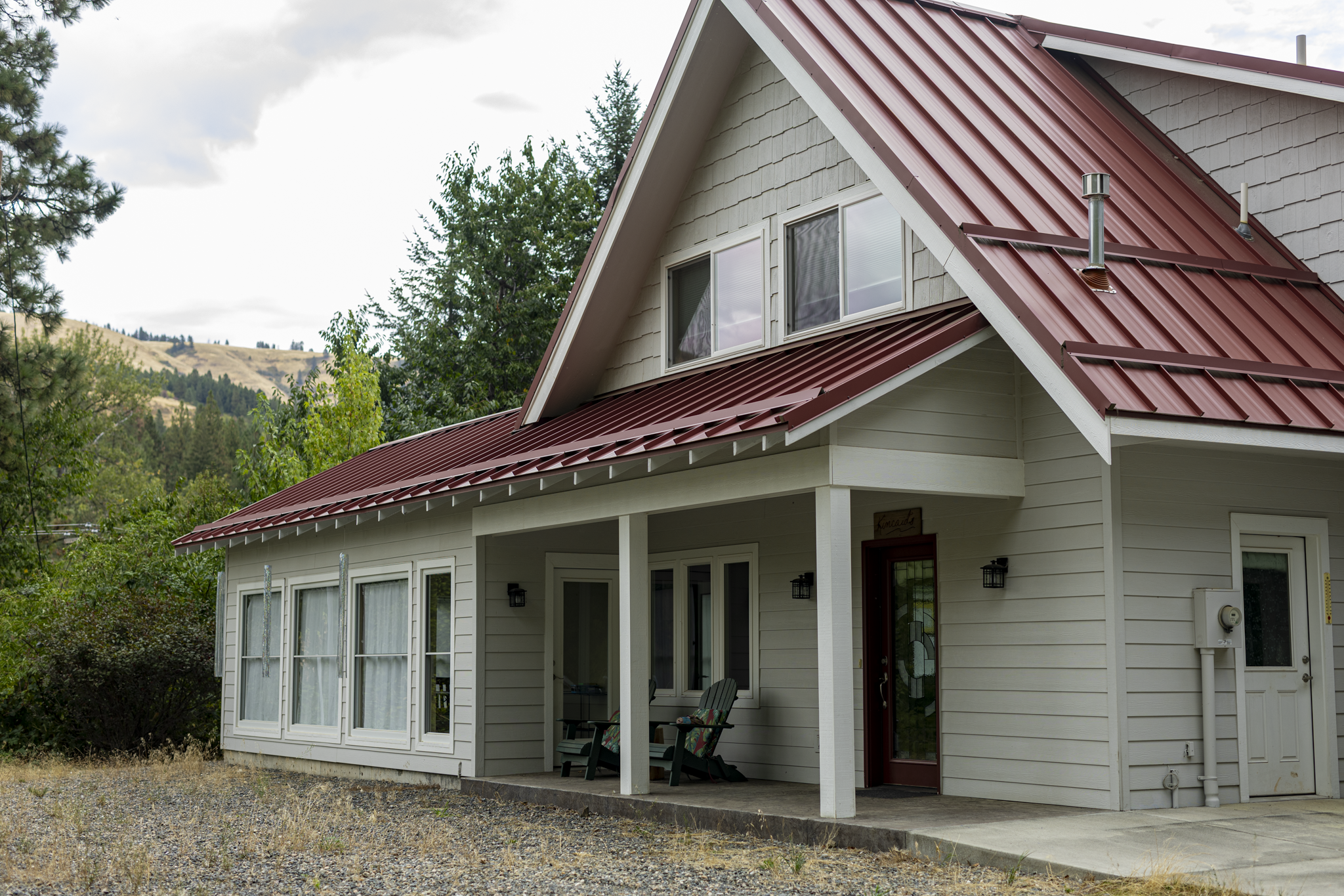 A two-story house with beige siding, large windows, and a red metal roof, situated in a rural area with trees and rolling hills in the background.