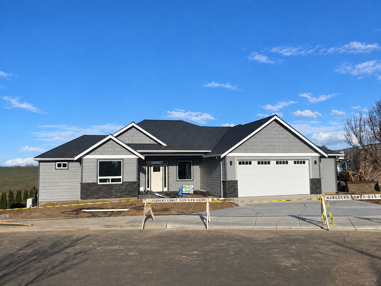 Newly constructed gray house with black roof and attached garage, building site with construction barriers and equipment, clear blue sky.