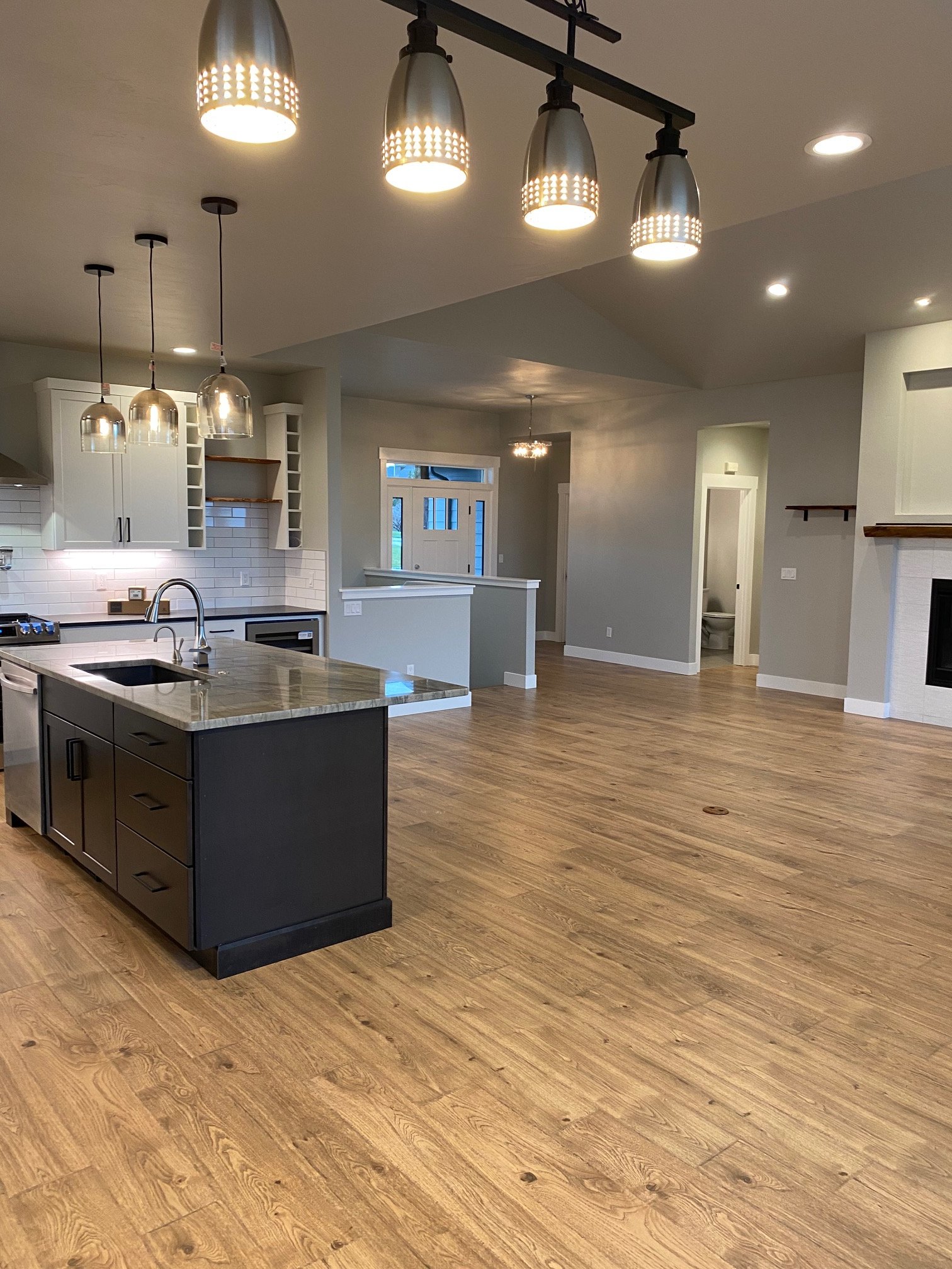 Modern open-concept kitchen and living area with wood flooring, white cabinets, pendant lights, and a small door leading to a bathroom.