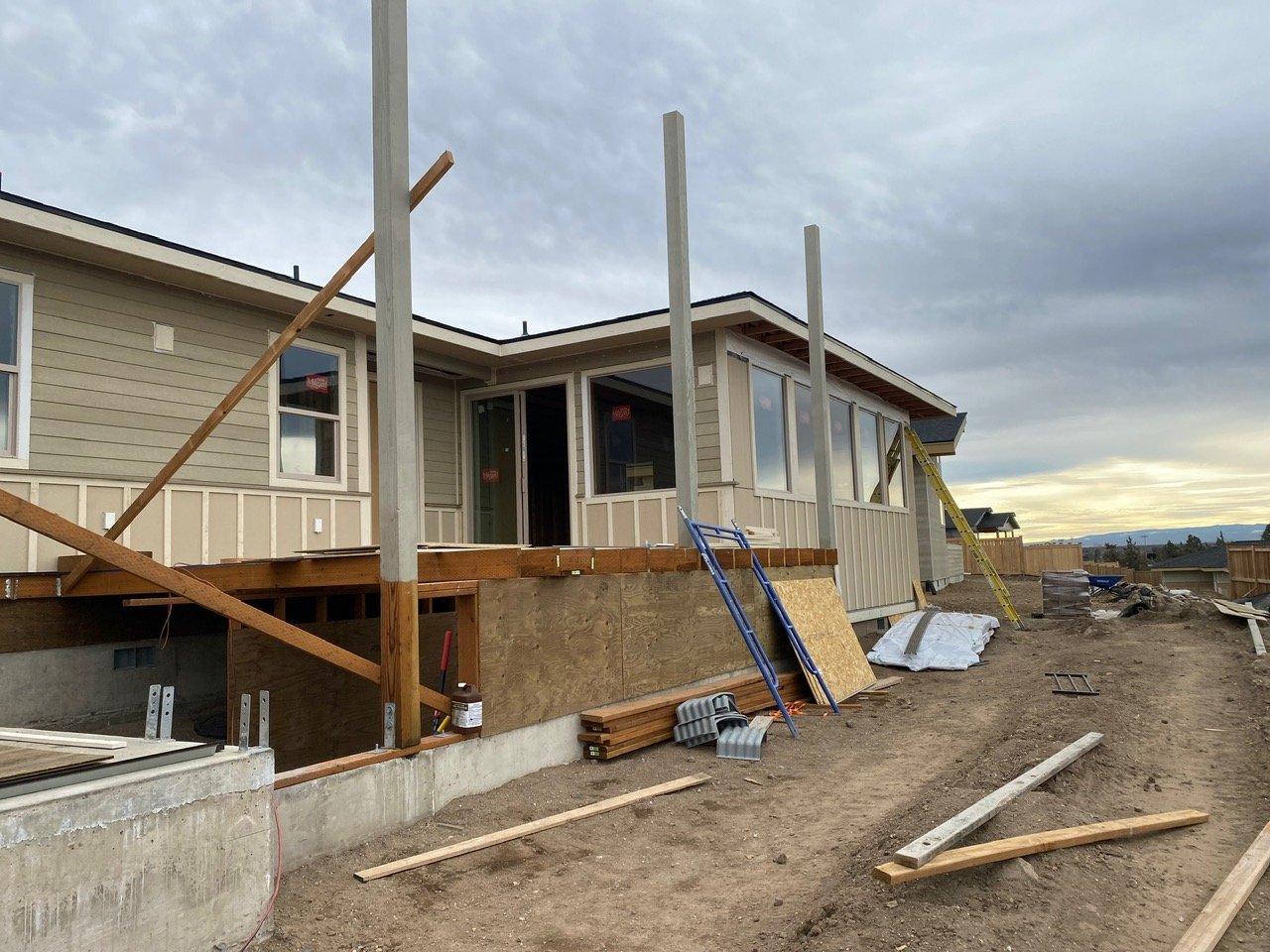 Construction site of a house with partially built wooden deck, ladders, and building materials, under overcast sky.