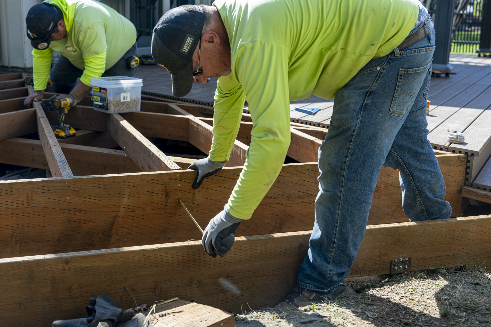 Two workers in neon yellow shirts and jeans are installing a wooden frame on an outdoor deck, using power tools and measuring tools.