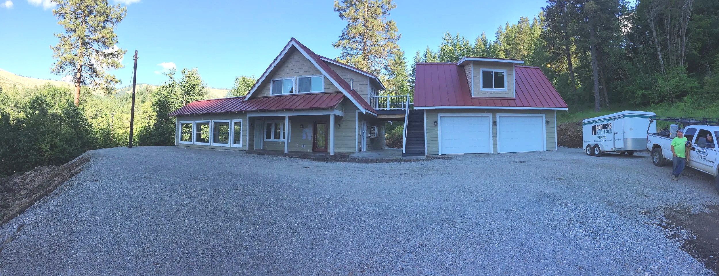 Newly constructed house with a red metal roof, multiple large windows, yellow siding, and a double garage, surrounded by trees and a gravel driveway, with a man standing near a moving truck.