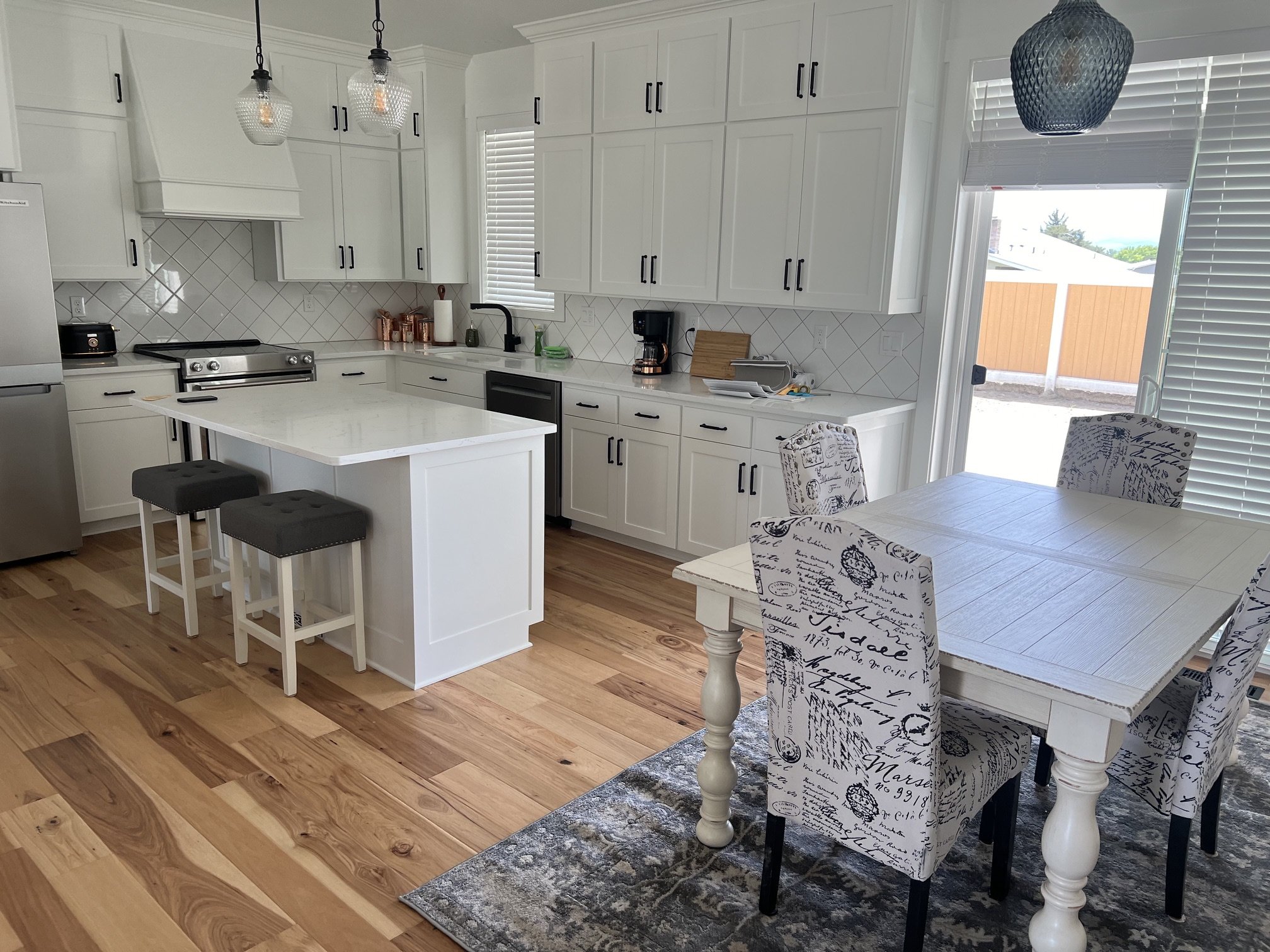Modern kitchen with white cabinets, black handles, and a white island counter with two gray stools. Features include a gas stove, a refrigerator, pendant lights, a black sink with a window, a coffee maker, copper containers, and a dining area with ch