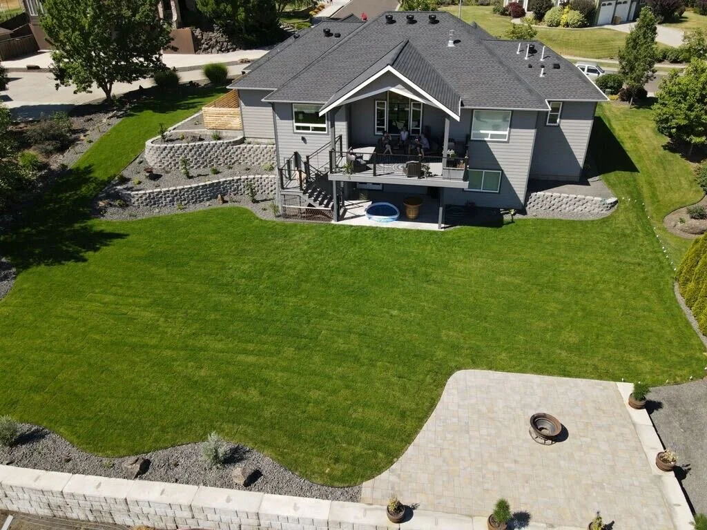 An aerial view of a house with a well-maintained backyard, including a green lawn, a paved patio area with a fire pit, and a tiered garden with stone retaining walls. The house has a covered deck with outdoor furniture.