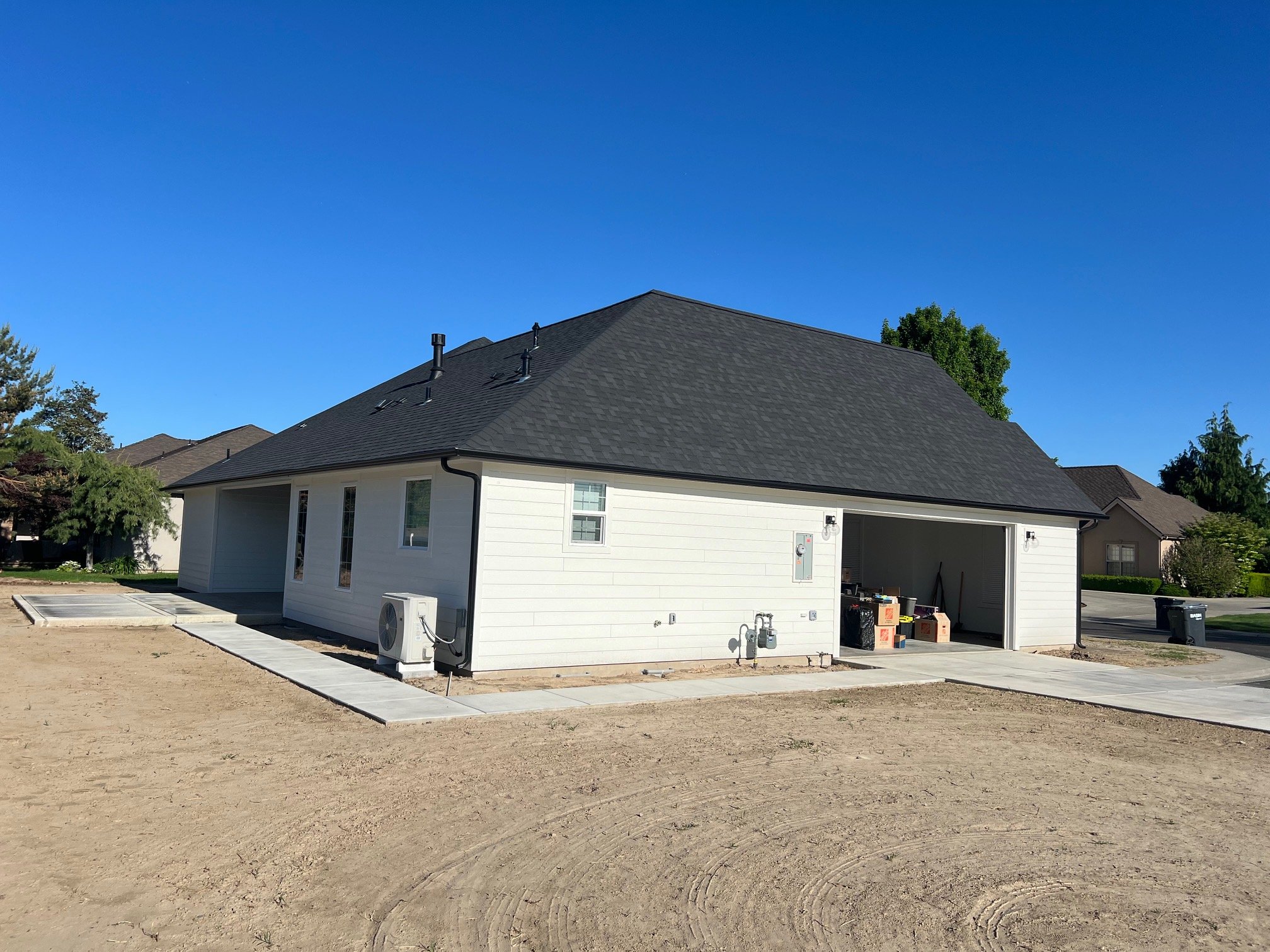 Side view of a white house with a dark gray roof, small windows, and a partially open garage, with a clear blue sky and trees in the background.