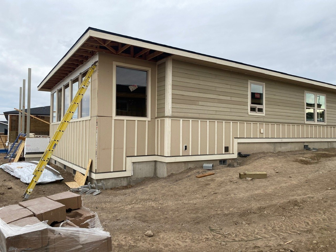 A house under construction with exterior siding partially installed, a yellow ladder leaning against the house, and construction materials on the ground. The house has multiple windows and is built on a raised foundation with dirt surrounding it.