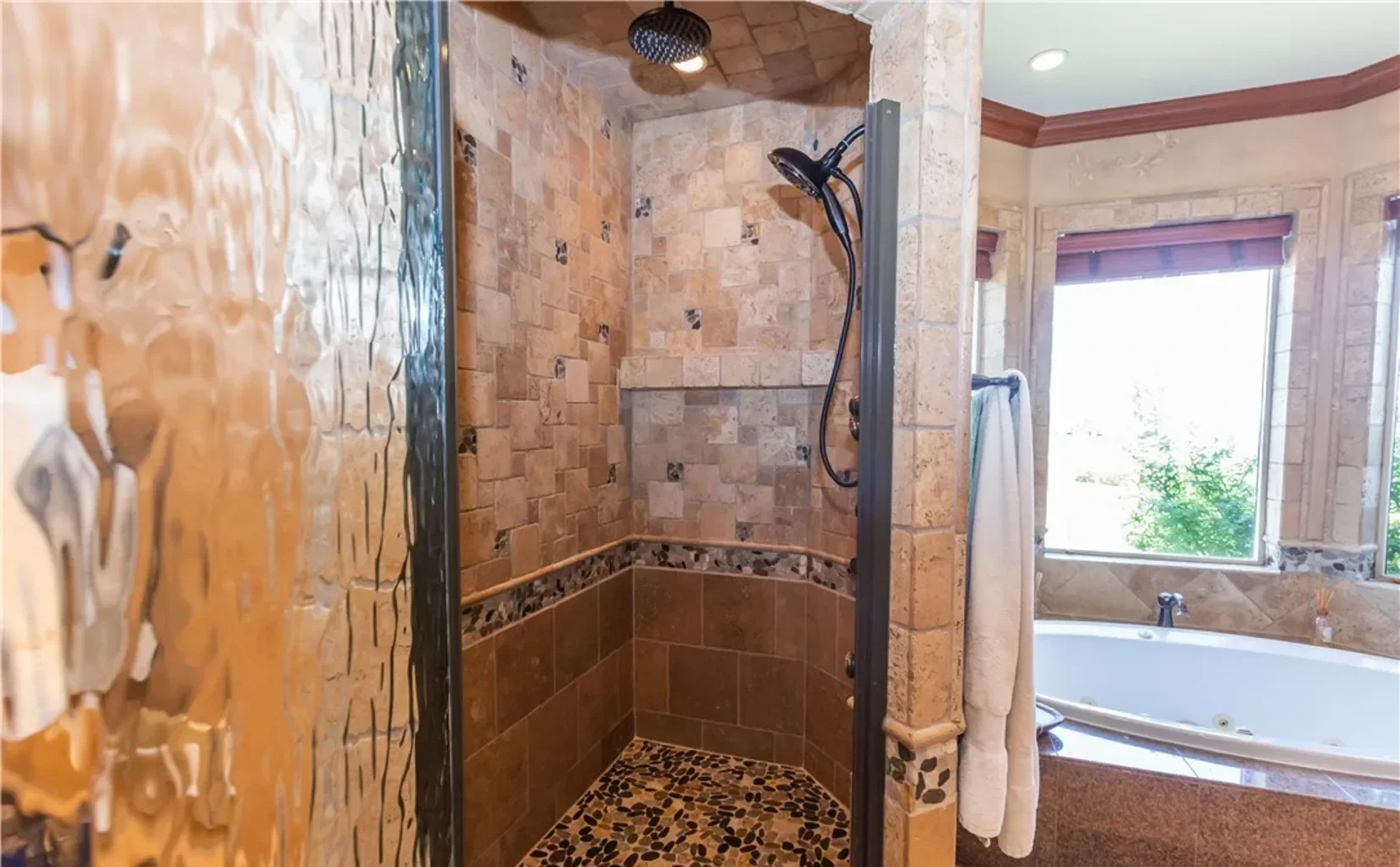 Bathroom with walk-in shower featuring tan and brown mosaic tile floor, beige tiled walls, and a shower head; adjacent is a bathtub with a window and a towel hanging on a rack.
