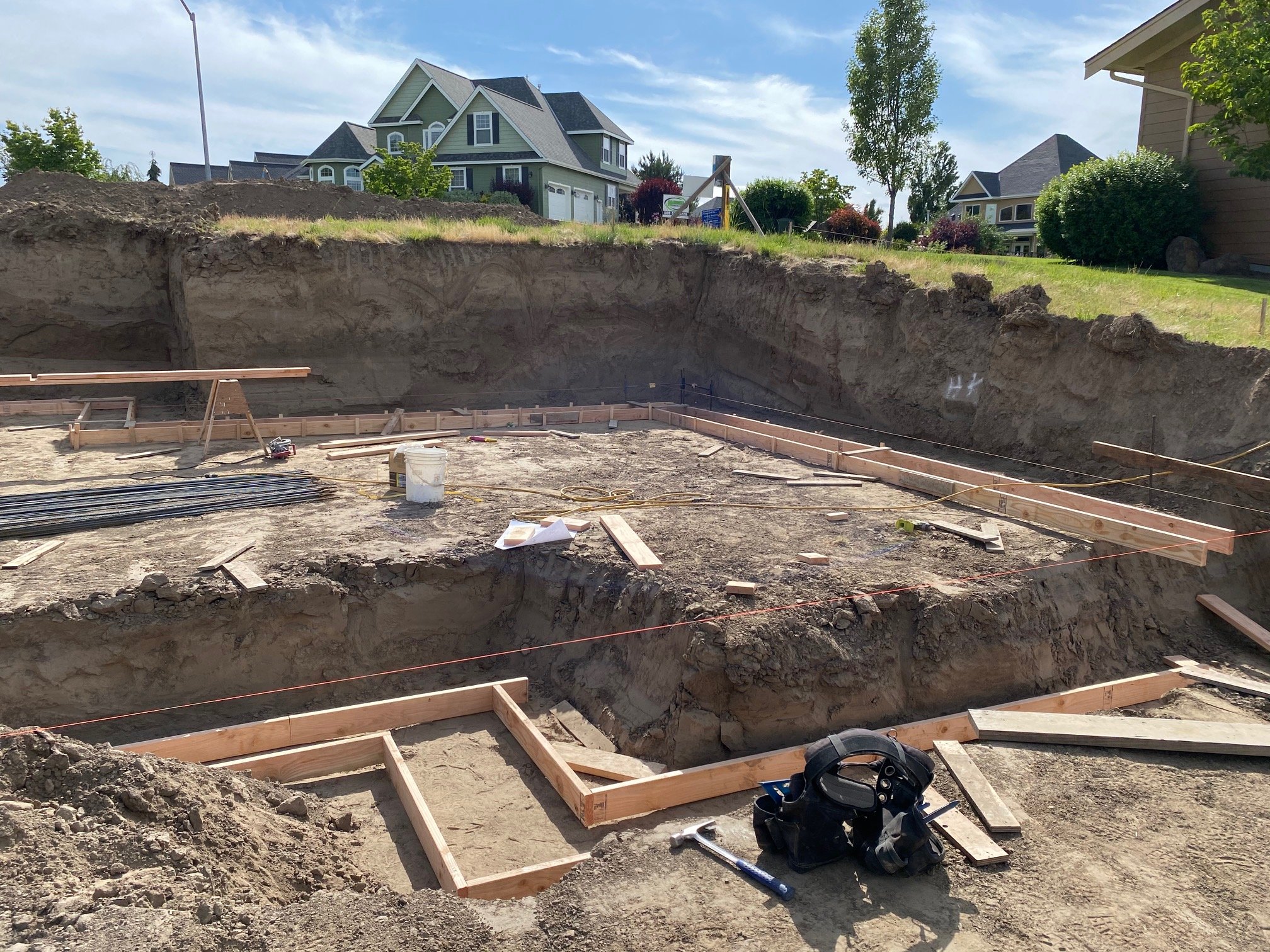 Construction site with wooden framing for building foundation, surrounded by dirt and construction tools, in a residential neighborhood with completed houses and trees in the background.
