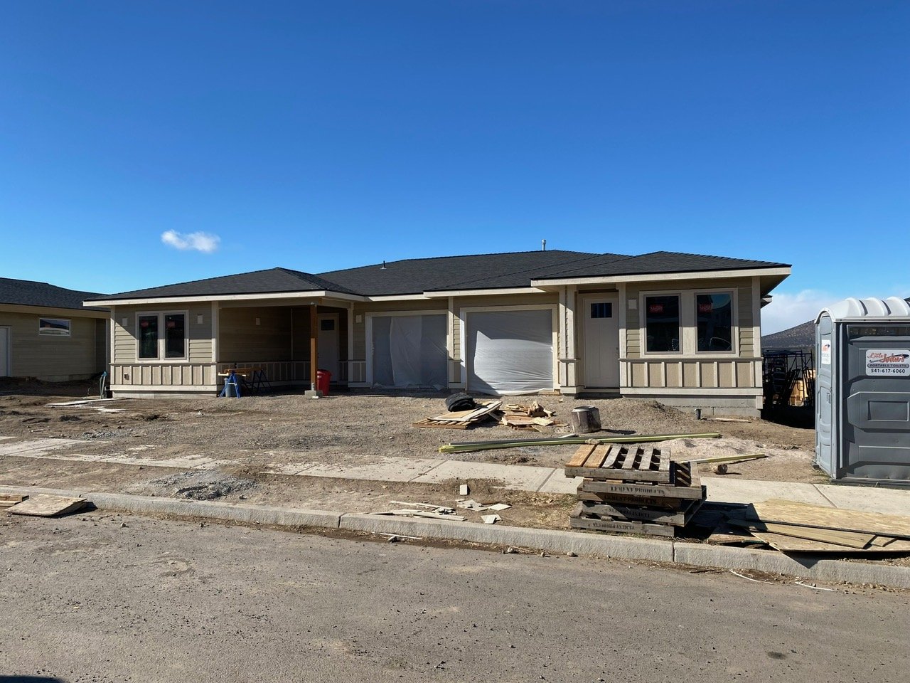 A house under construction with exterior walls and roof completed, but driveway and yard still unfinished, construction materials, pallets, and a portable toilet in the yard.