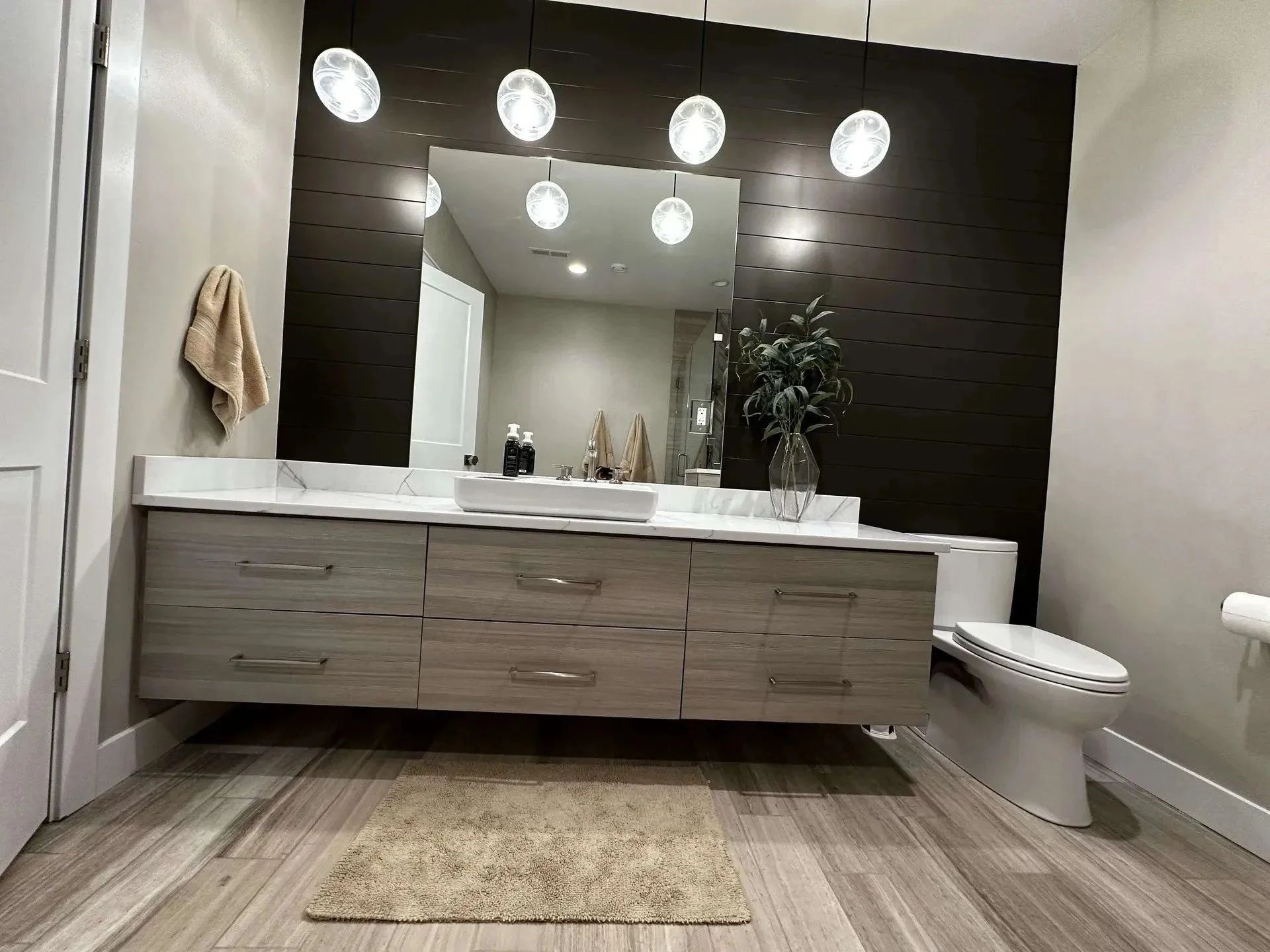 Modern bathroom with a large mirror, dark wood accent wall, white countertop with sink, beige towel on hook, decorative plant in a glass vase, bottles of soap or lotion, toilet with closed lid, overhead lighting fixtures, wood-look tiled floor, and a