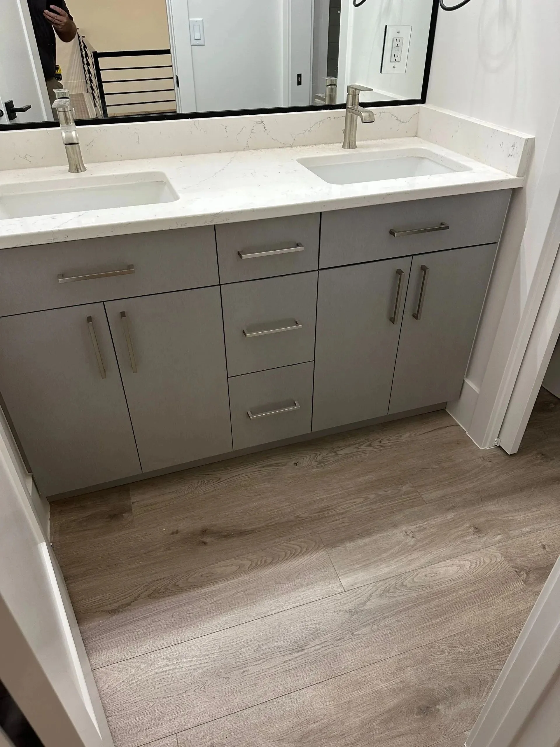A bathroom vanity with a white countertop and two sinks, each with a modern faucet. There is a large mirror above the vanity, and below are multiple drawers and cabinets. The flooring is wood-style vinyl.