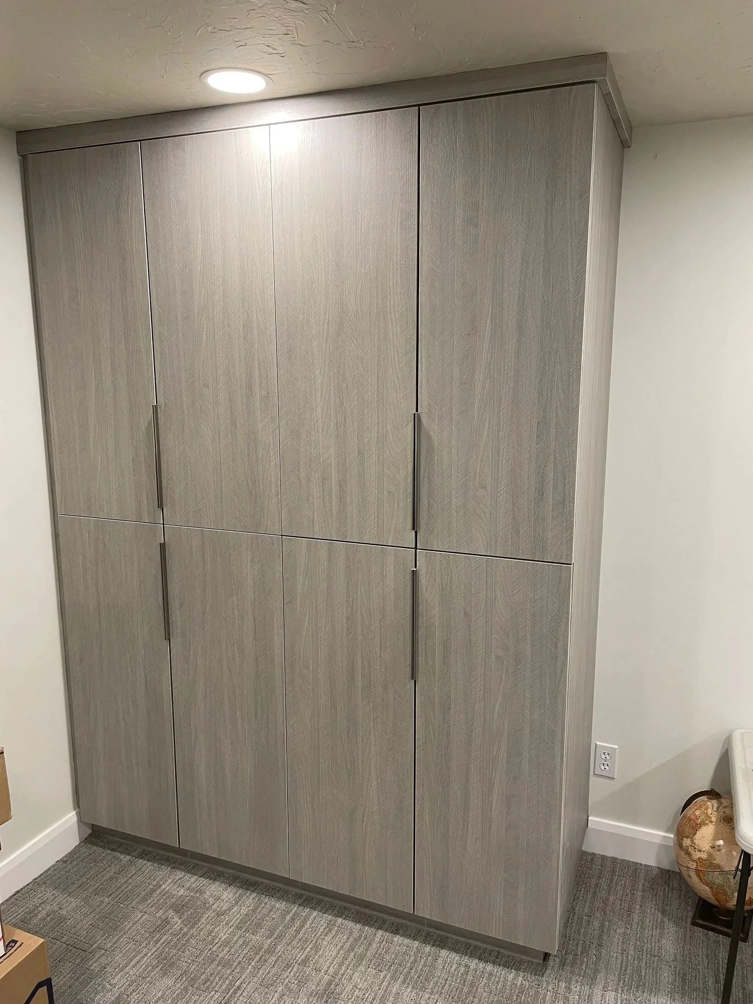 A modern corner wardrobe with four beige doors in a room with neutral walls and carpeting, a potted globe on a small table next to the wardrobe, and a box in the bottom left corner.