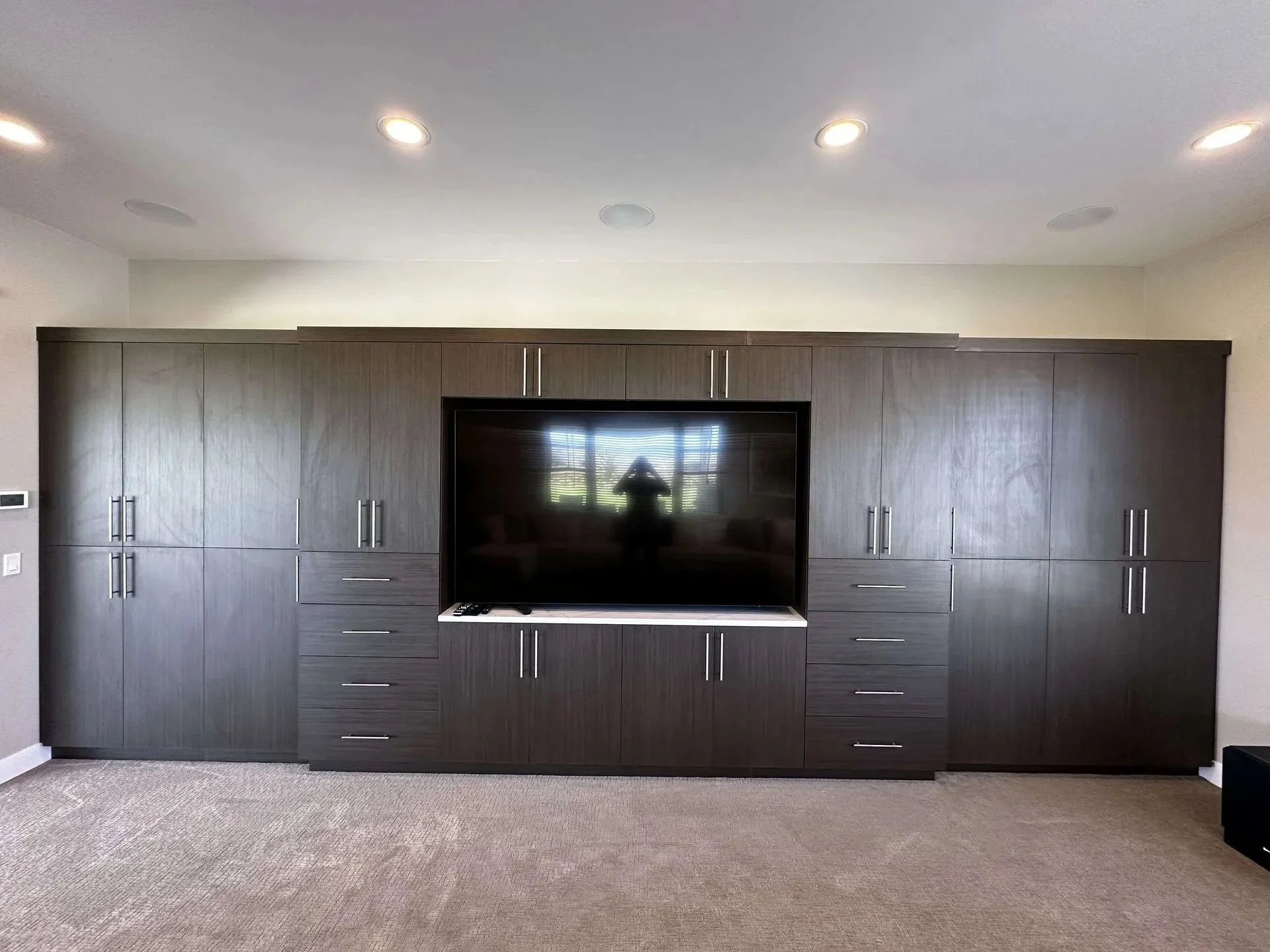 A large built-in dark wood entertainment center with multiple cabinets and drawers surrounding a flat-screen television, set against a white wall with a beige carpeted floor.