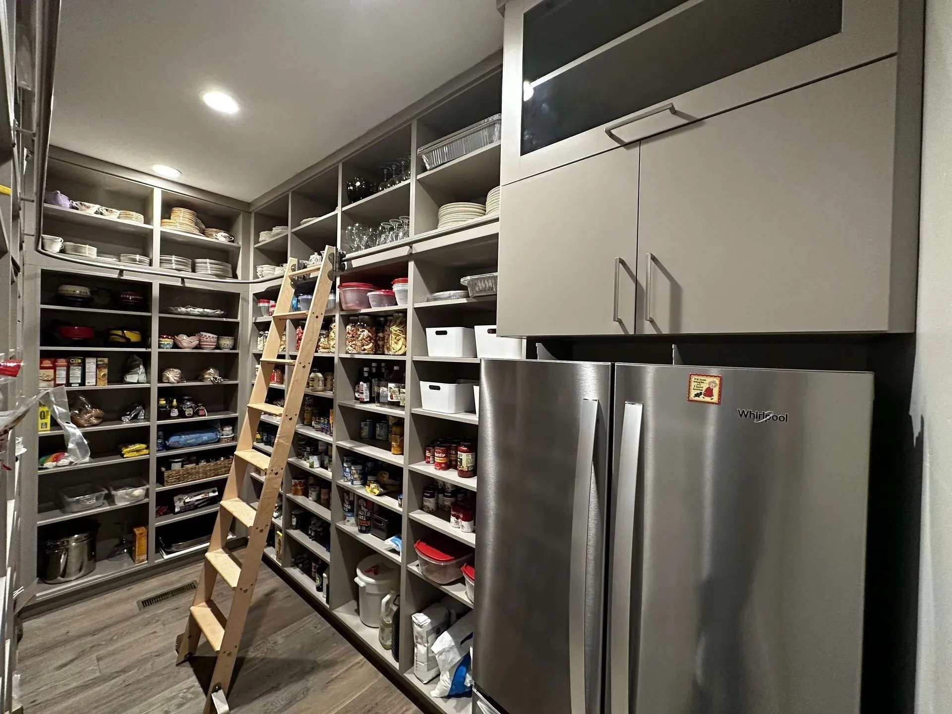A walk-in pantry with shelves filled with dishes, jars, and pantry items, a step ladder, and a side-by-side refrigerator.