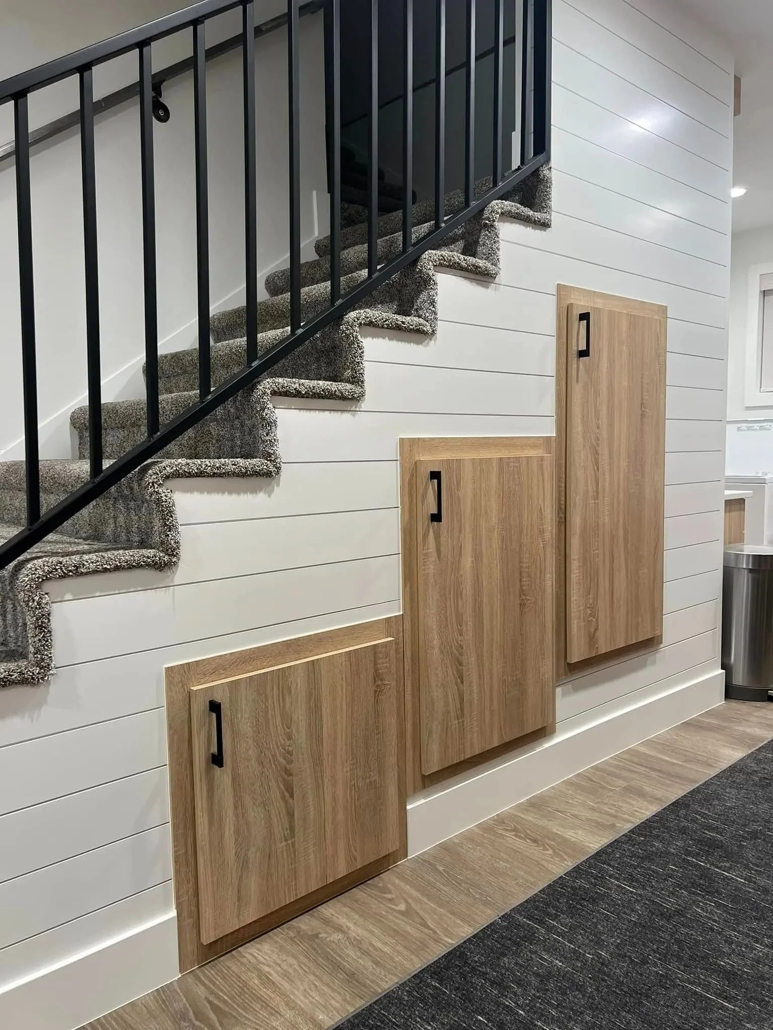 Interior view of a staircase with carpeted steps, black metal railing, white shiplap wall, wooden cabinets, and a trash can at the bottom right corner.