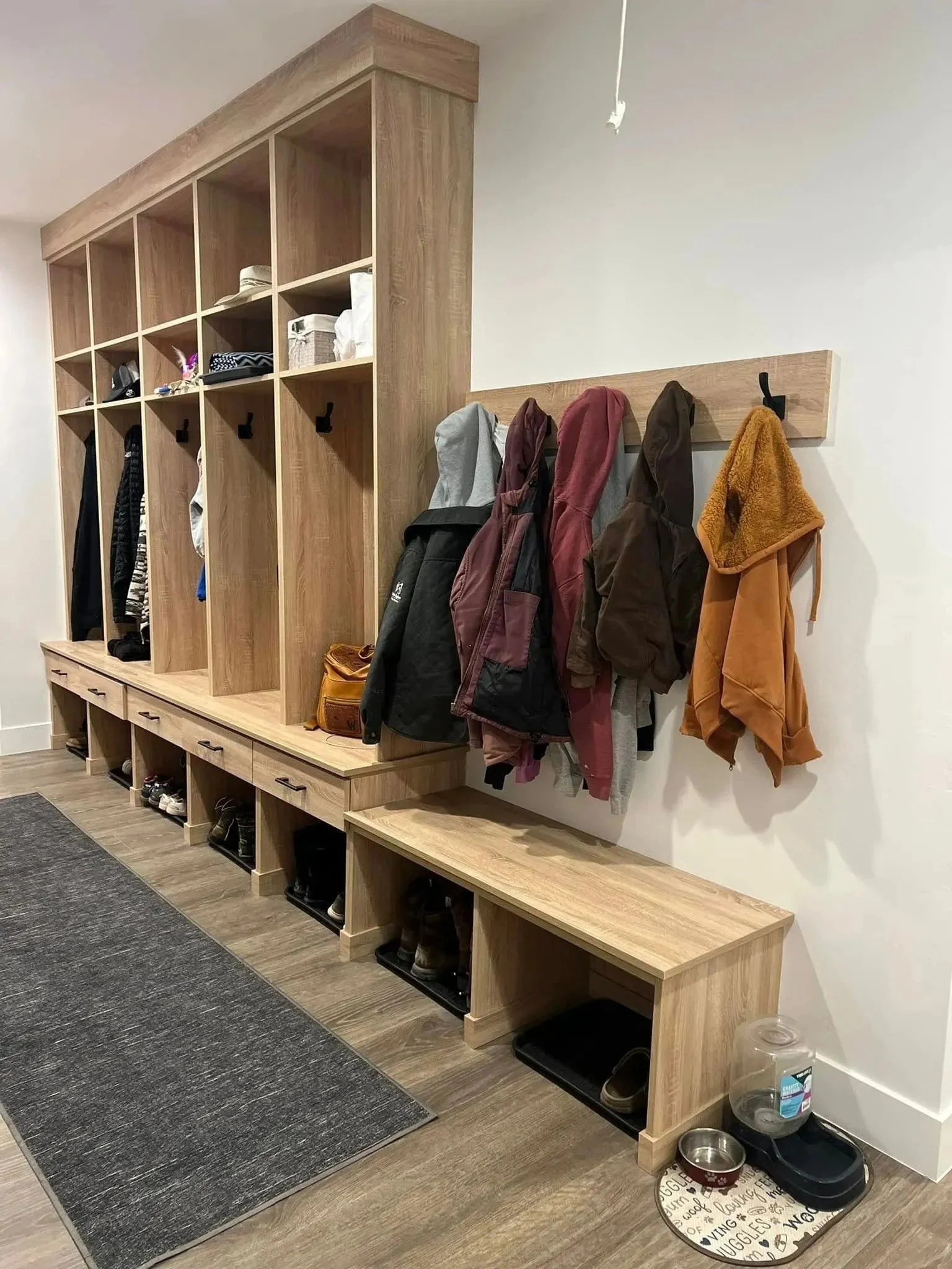 Wooden hall tree with hooks and cubbies for shoes, containing jackets, hats, and hats, next to a white wall with a small pet water and food station on the floor.