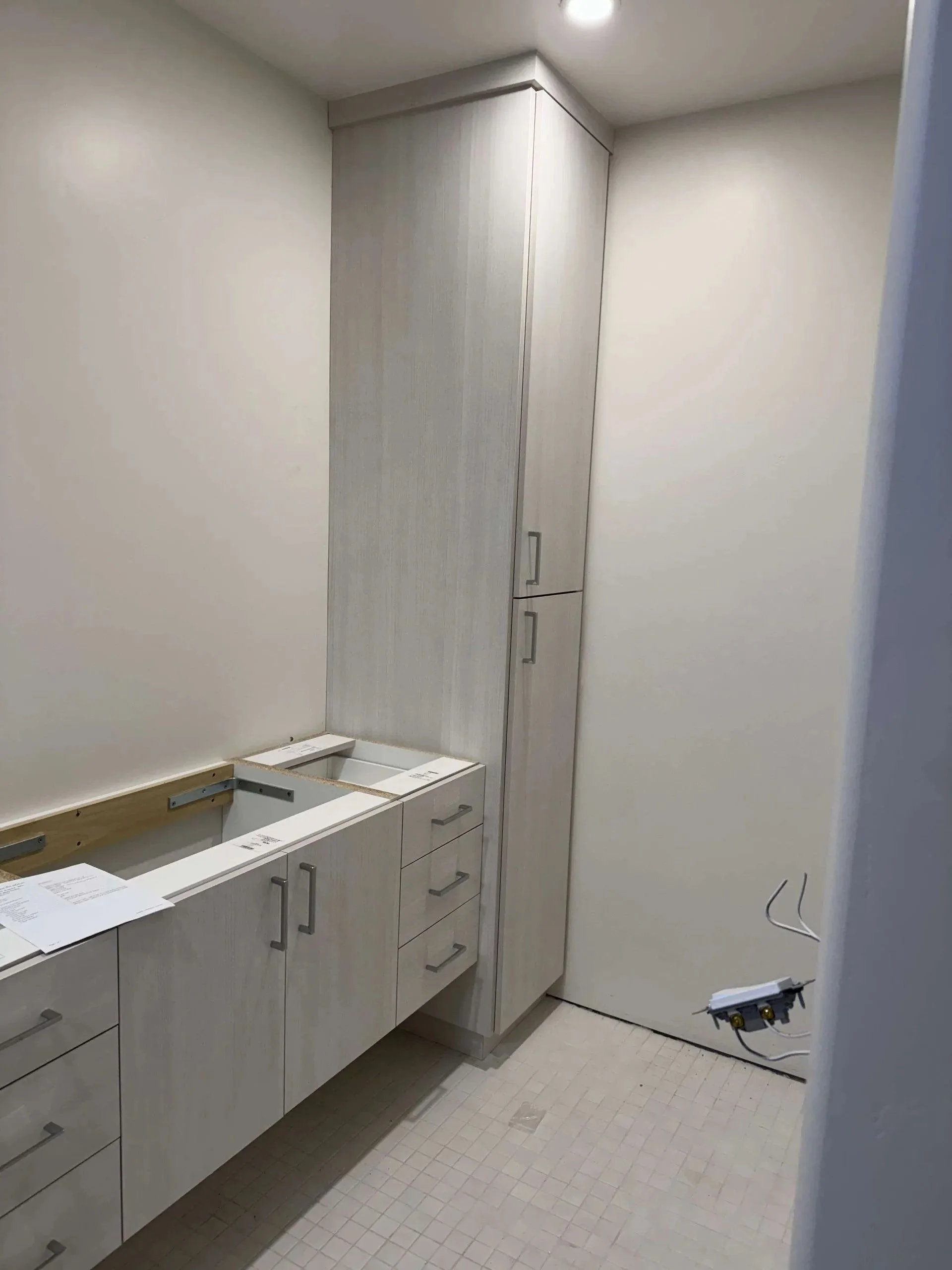 An unfinished bathroom with white cabinets and a tiled floor, with a section of countertop missing and exposed wiring.