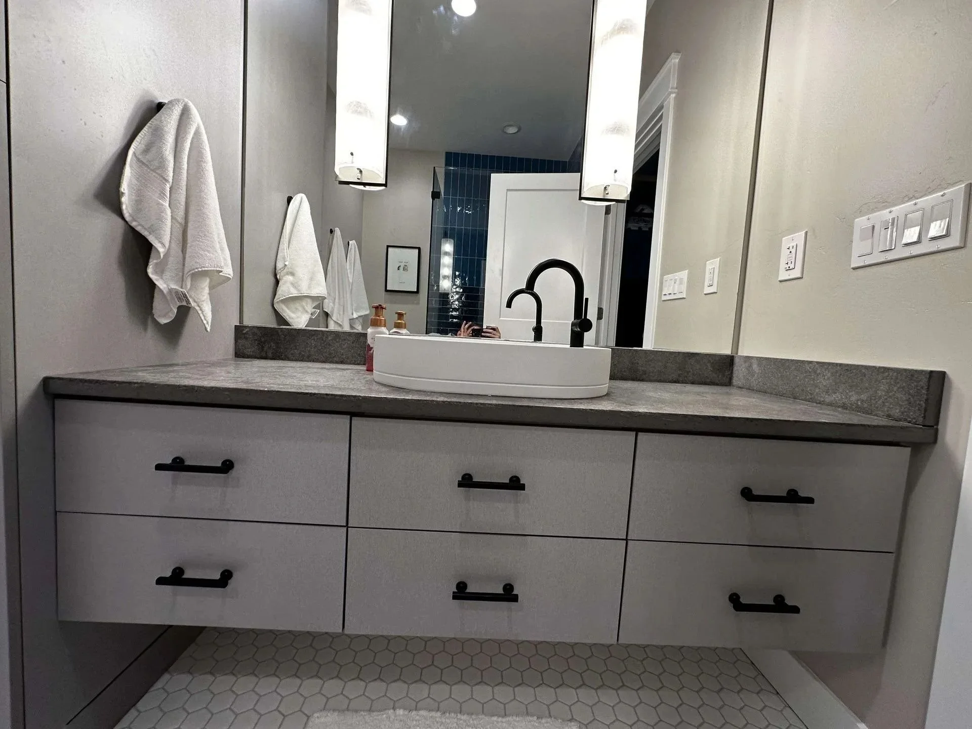 Bathroom vanity with white cabinets, black handles, gray countertop, and a white vessel sink with a black faucet. Towels hang on the wall, and a large mirror above reflects the room. The bathroom has a hexagonal tile floor and a door leading to anoth