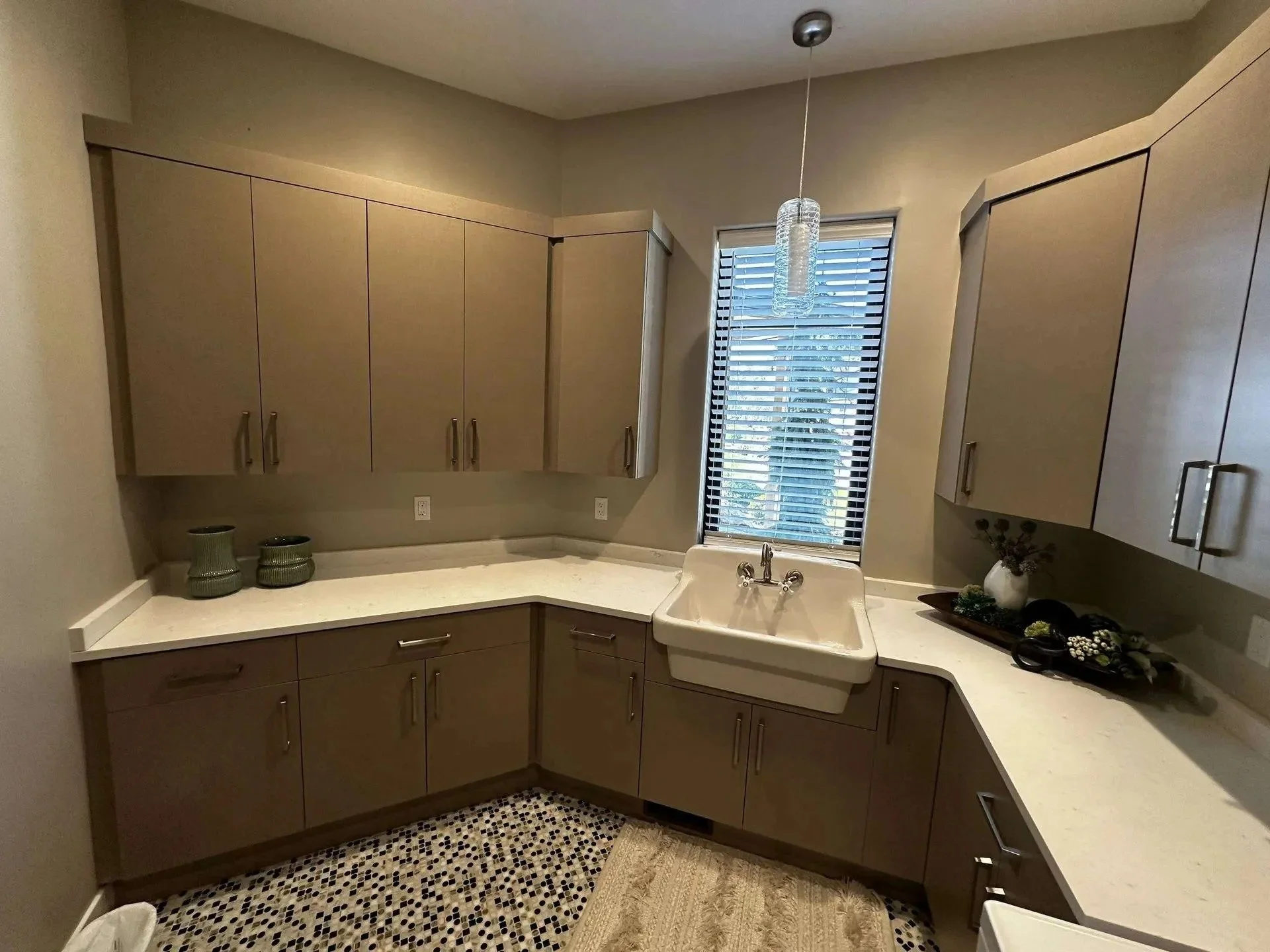 Modern kitchen with beige cabinets, white countertops, a white sink, a window with blinds, decorative items including vases and plants, and a small tiled floor area.