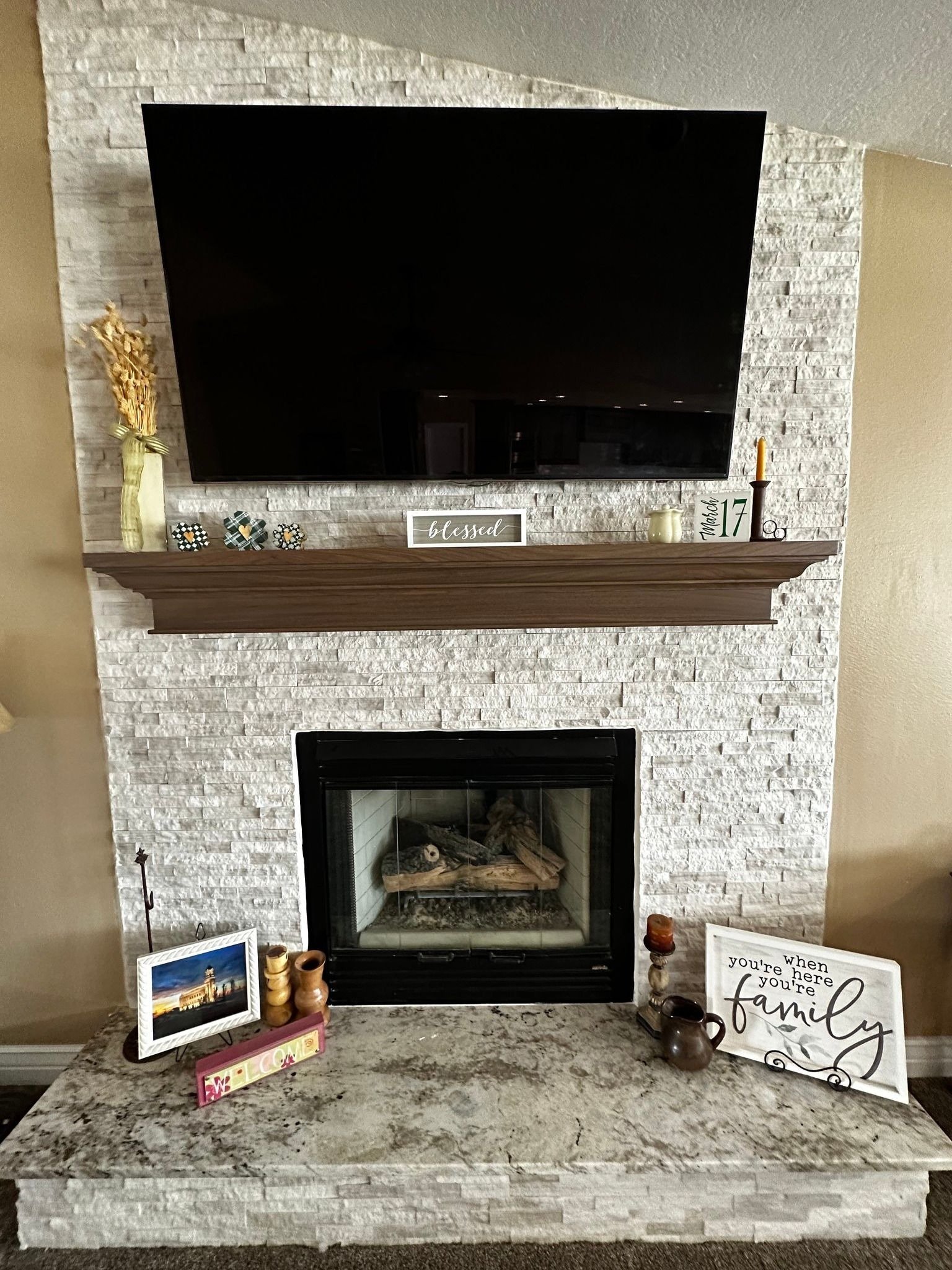 Fireplace with white decorative brick surround, wooden mantle, a large flat-screen TV mounted above, and various decorative items on the mantle and surrounding area including a framed picture, a sign that says 'blessed,' a calendar, a small jar, and 