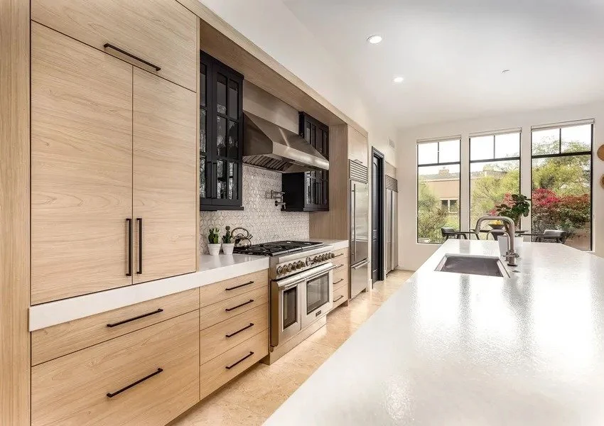 Modern kitchen with light wood cabinets, stainless steel appliances, black upper cabinets, white countertops, and large windows with outdoor view.